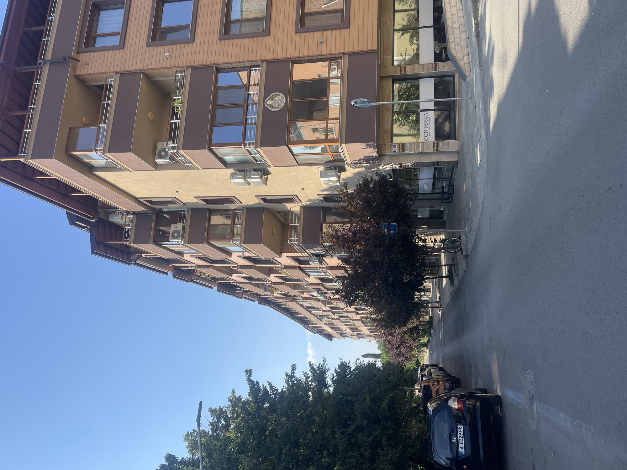 The image depicts a street scene in Burgas, Bulgaria, on a clear day with a bright blue sky. On the right side of the frame, a multi-story residential building with brown and beige facades dominates the view. The building features numerous balconies with modern railings, air conditioning units, and large windows. The ground floor of the building houses a commercial establishment with the name "CONTESSA" visible on its facade, indicating a beauty salon or academy.
On the left side of the street, a row of parked cars lines the asphalt road. The closest car is a black hatchback with a license plate reading "A 0229 PK". Further down the street, more vehicles are parked, and deciduous trees with dark red foliage create a canopy over the sidewalk. In the distance, the street appears to continue between buildings and trees. A bicycle is visible parked on the right sidewalk. The environment suggests a residential and commercial area within a city.