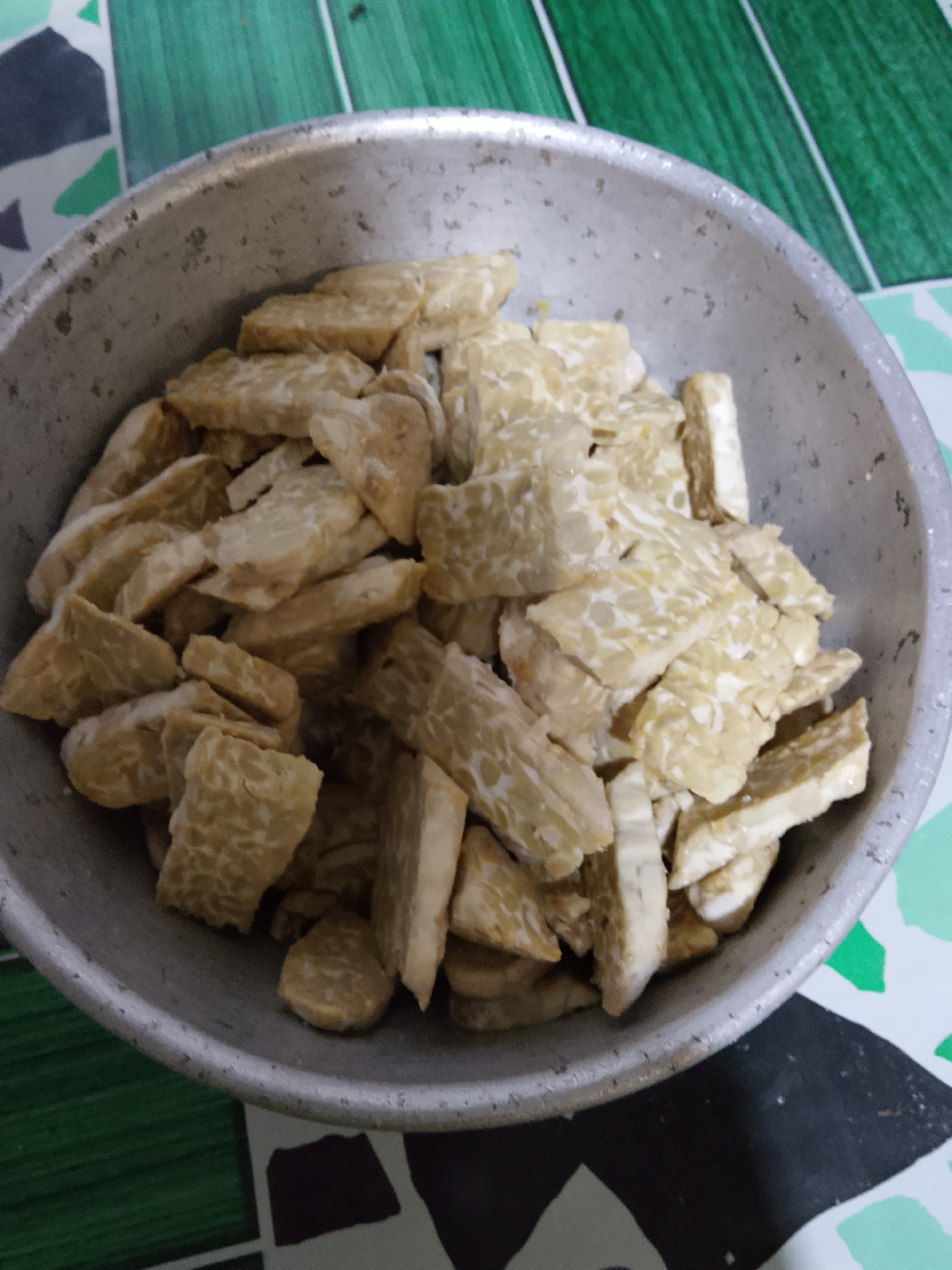 In Merbopuntong, Indonesia, a close-up, overhead shot shows a metal bowl filled with sliced tempeh. The tempeh is cut into irregular, bite-sized pieces and appears to be raw or unseasoned. The bowl is placed on a surface with a geometric pattern of green, white, and black. The lighting is dim, casting soft shadows.