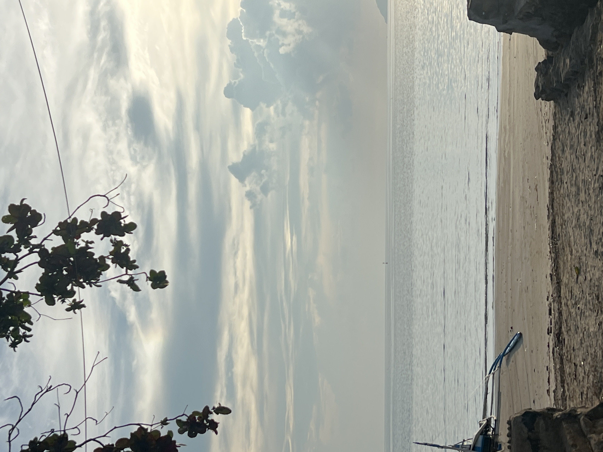 This image captures a serene beach scene in Cuyo, Philippines. The foreground shows a sandy beach with some debris and a partially visible structure made of rocks and concrete on the right. A small, blue and white outrigger boat rests on the sand near the waterline, its long outriggers extending outwards. The sea is calm and stretches to the horizon, where a small marker buoy is visible in the distance. The sky is partly cloudy, with wispy cirrus clouds and larger cumulus formations, illuminated by soft, diffused light, suggesting either early morning or late afternoon. Dark green foliage hangs from branches in the upper left corner of the frame, framing the sky and the sea.