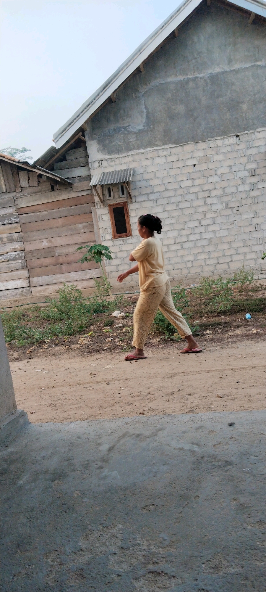 In Tanjung Ratu Ilir, Indonesia, a young woman wearing a yellow patterned pajama set and pink flip-flops walks on a dirt path next to a brick building with a corrugated metal roof over a small window. To the left of the brick building is a wooden structure with horizontal planks. Vegetation grows along the ground, and a young tree is visible. The sky is overcast.