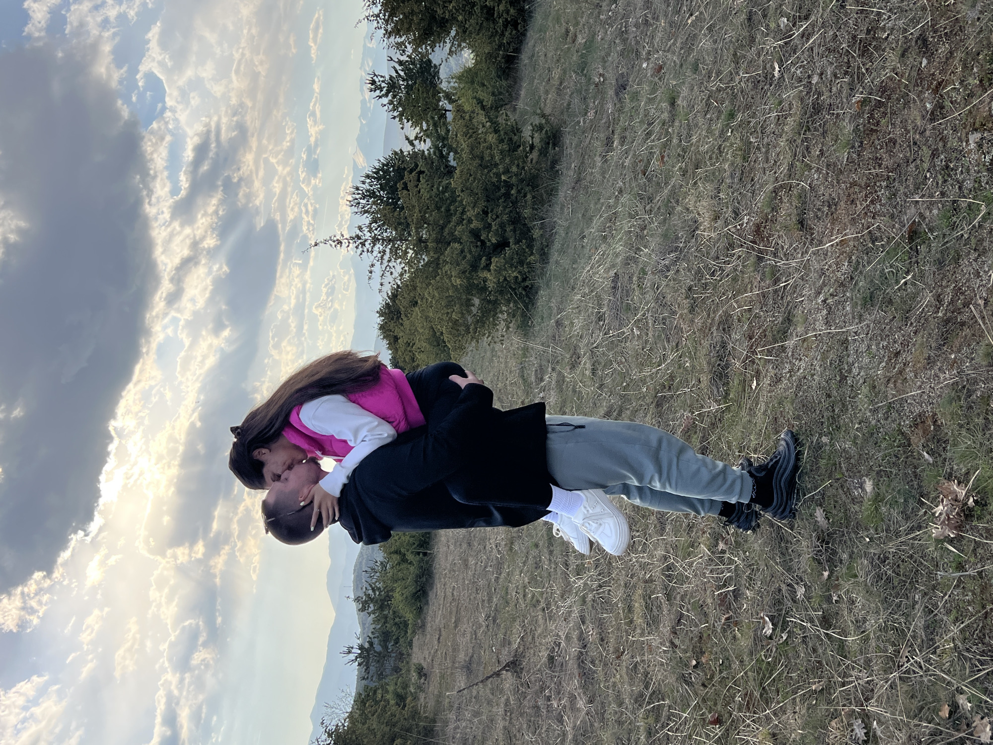 A couple is embracing in an outdoor setting. The woman has long brown hair and wears a pink vest over a white shirt. The man is carrying her, wearing a black jacket and grey pants. They are kissing. The background includes a grassy hillside, trees, mountains, and a cloudy sky. The location appears to be on a hillside in Kribul, Bulgaria.