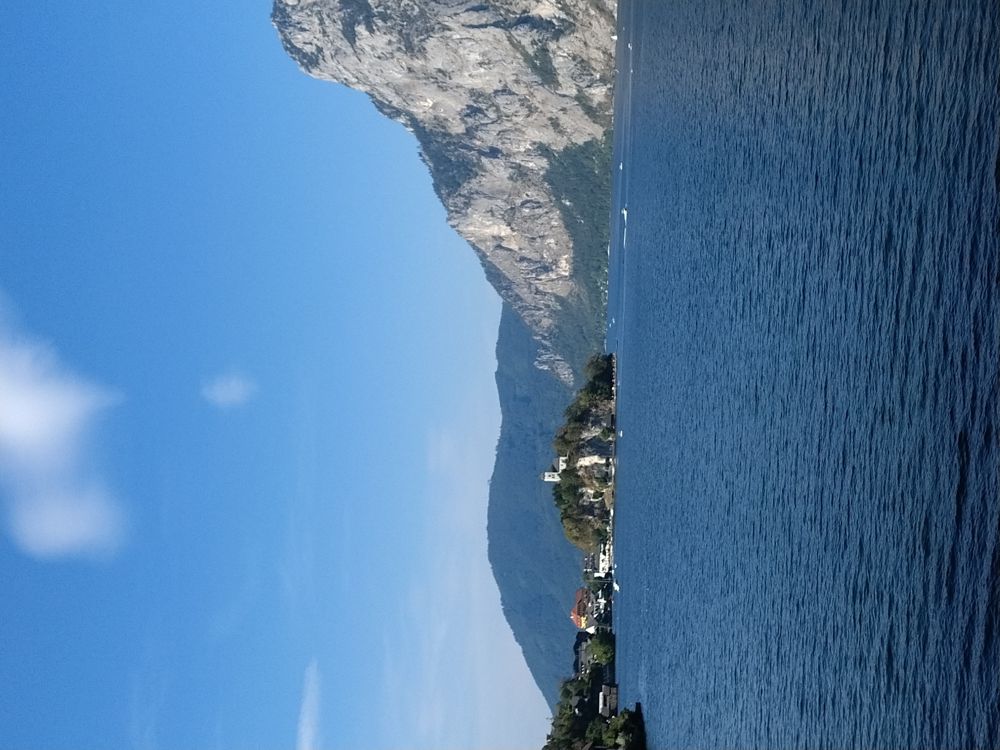 The image depicts a scenic view of Traunkirchen, Austria. In the foreground, a large expanse of deep blue water, likely Lake Traun, stretches across the frame. The water's surface is gently rippled, indicating a calm day. In the mid-ground, a picturesque village is situated on the shore and extends onto a small island. The island is crowned by a white church with a prominent steeple, nestled amongst lush green trees. Adjacent to the island, houses with red and yellow roofs are visible along the waterfront, suggesting a charming European village. Several white boats are dotted on the water near the village. Behind the village, rolling green hills rise, covered in dense forest. The background is dominated by the imposing, rugged face of a large, grey, rocky mountain. The sky above is a clear, bright blue with a few wispy white clouds. The overall scene is one of natural beauty and tranquility, showcasing the harmonious blend of a village, a lake, and mountainous terrain.