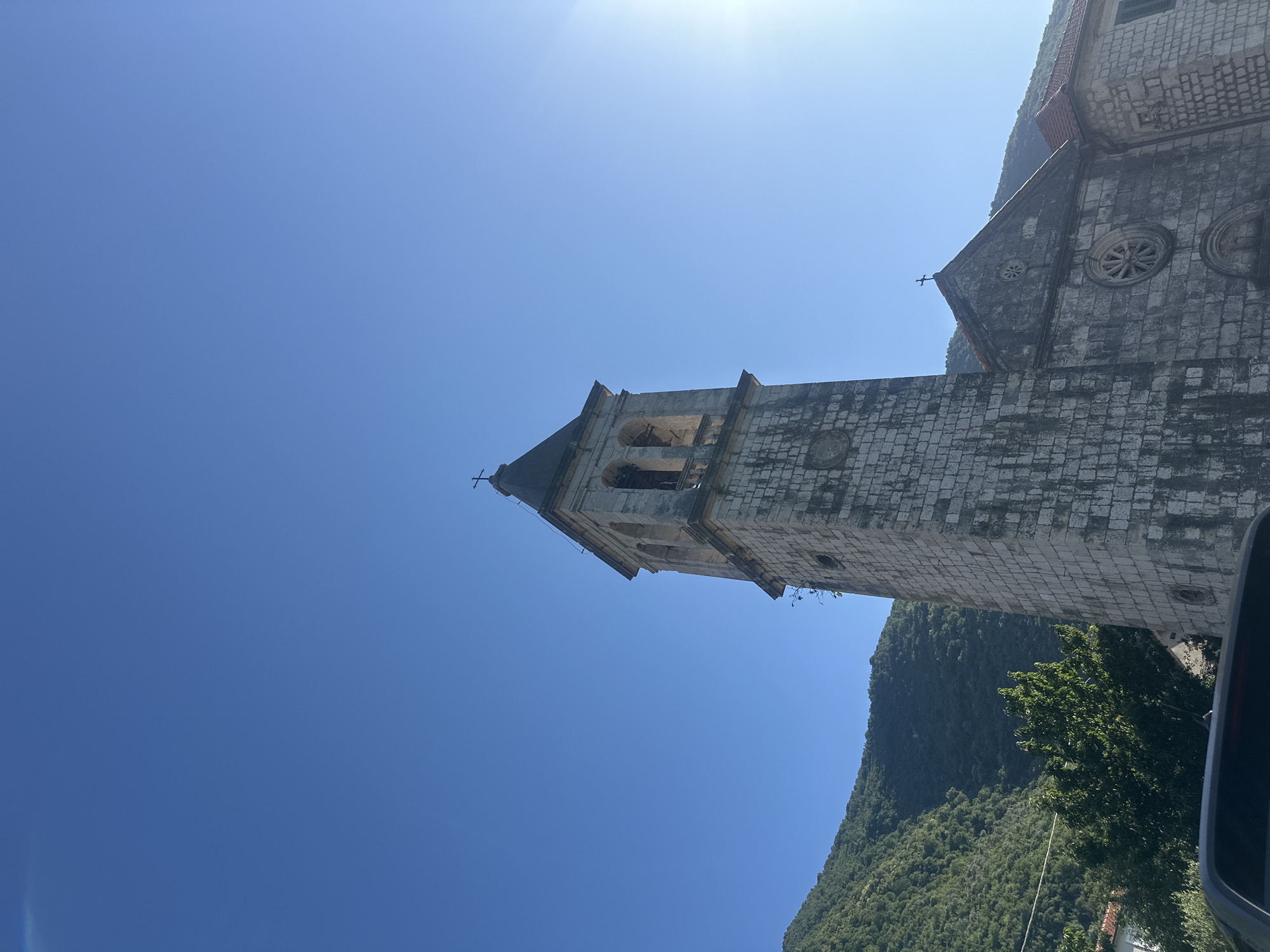 This image depicts the Church of the Nativity of the Blessed Virgin Mary in Prčanj, Montenegro, under a clear blue sky on a sunny day. The central subject is the church's prominent bell tower, constructed from weathered grey stone blocks. The tower is square-shaped, featuring arched belfry openings near its top and a pyramidal dark-colored roof capped with a cross. Below the belfry, a circular indentation is visible on the stone facade. To the right of the bell tower, a portion of the main church building is visible, also constructed from similar stone blocks. This section displays a gabled roof with a cross at its peak and two circular windows, one larger (resembling a rose window) below the gable and a smaller one above it. An adjacent stone building with a terracotta tiled roof is partially visible to the far right. In the background, lush, densely forested green mountains rise under a brilliant, cloudless blue sky. A strong lens flare or sun glare is evident in the upper right quadrant of the sky. The very bottom of the frame shows a dark-colored vehicle. No individuals are present in the scene.