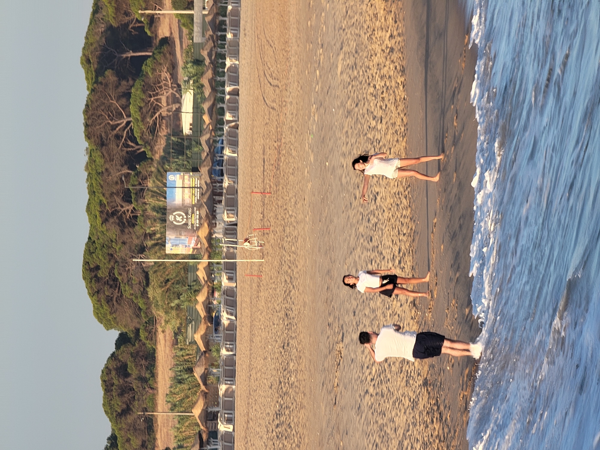 This media file depicts a beach scene in Quarteira, Portugal. The image features three individuals on a sandy beach, with the ocean visible on the right.
The first person, a woman, is lying on her back on the sand, wearing a white top and shorts. The second person, also a woman, is standing and appears to be looking towards the ocean, dressed in a white top and shorts. The third person, a man, is lying on his back on the sand, wearing a white t-shirt and dark shorts.
The setting is a beach with sand dunes and pine trees in the background on the left. Rows of sun loungers and umbrellas are visible in the middle ground. The ocean on the right has white waves breaking on the shore.
The time of day appears to be late afternoon or early evening, as suggested by the warm, soft lighting and the long shadows cast on the sand. The weather seems to be pleasant, with clear skies.
There is visible text on a billboard or sign in the background on the left, but it is too small and blurry to be read.