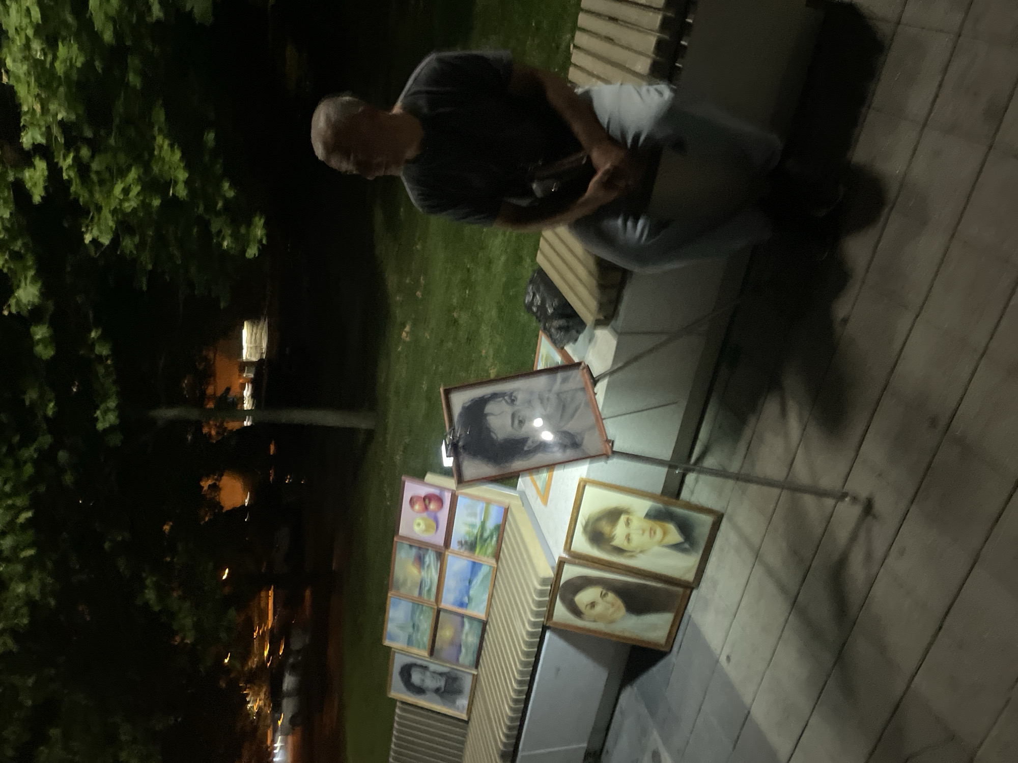 A man sits on a park bench at night, with several framed paintings displayed on the bench and a nearby easel. The paintings include portraits, landscapes, and still lifes of fruit. The man appears to be selling his artwork. The background shows trees and distant city lights, suggesting an urban park setting. The overall lighting is dim, consistent with nighttime. The location of Sadovyi, Russia is not discernible from the image itself.
