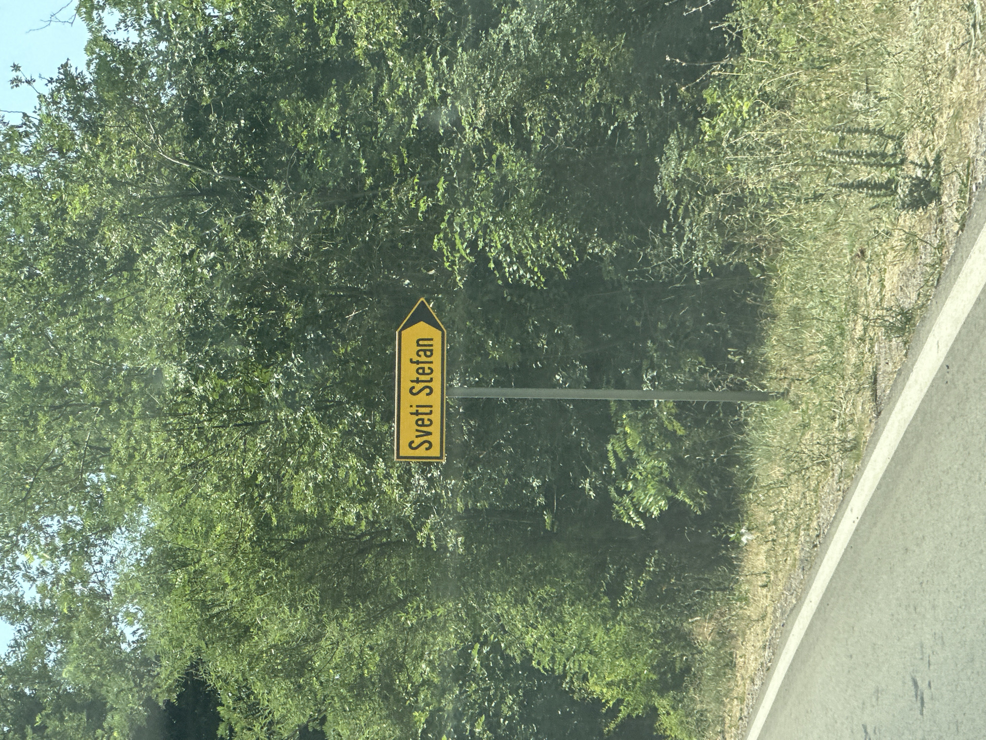 The image shows a yellow directional sign with black text in front of a dense green forest. The sign is mounted on a metal pole. The text on the sign reads "Sveti Stefan" in a stylized font. The sign is oriented vertically and appears to be pointing towards the left of the frame. The forest is lush and leafy, with sunlight filtering through the trees, suggesting a daytime scene. The background is dominated by the greenery of the forest, with a glimpse of a paved road on the right side of the image. The overall impression is that of a road trip or a scenic drive, likely in the vicinity of Sveti Stefan, Montenegro, as indicated by the text on the sign. The weather appears to be clear and sunny. No people or specific activities are depicted, focusing solely on the road sign and the natural surroundings.
