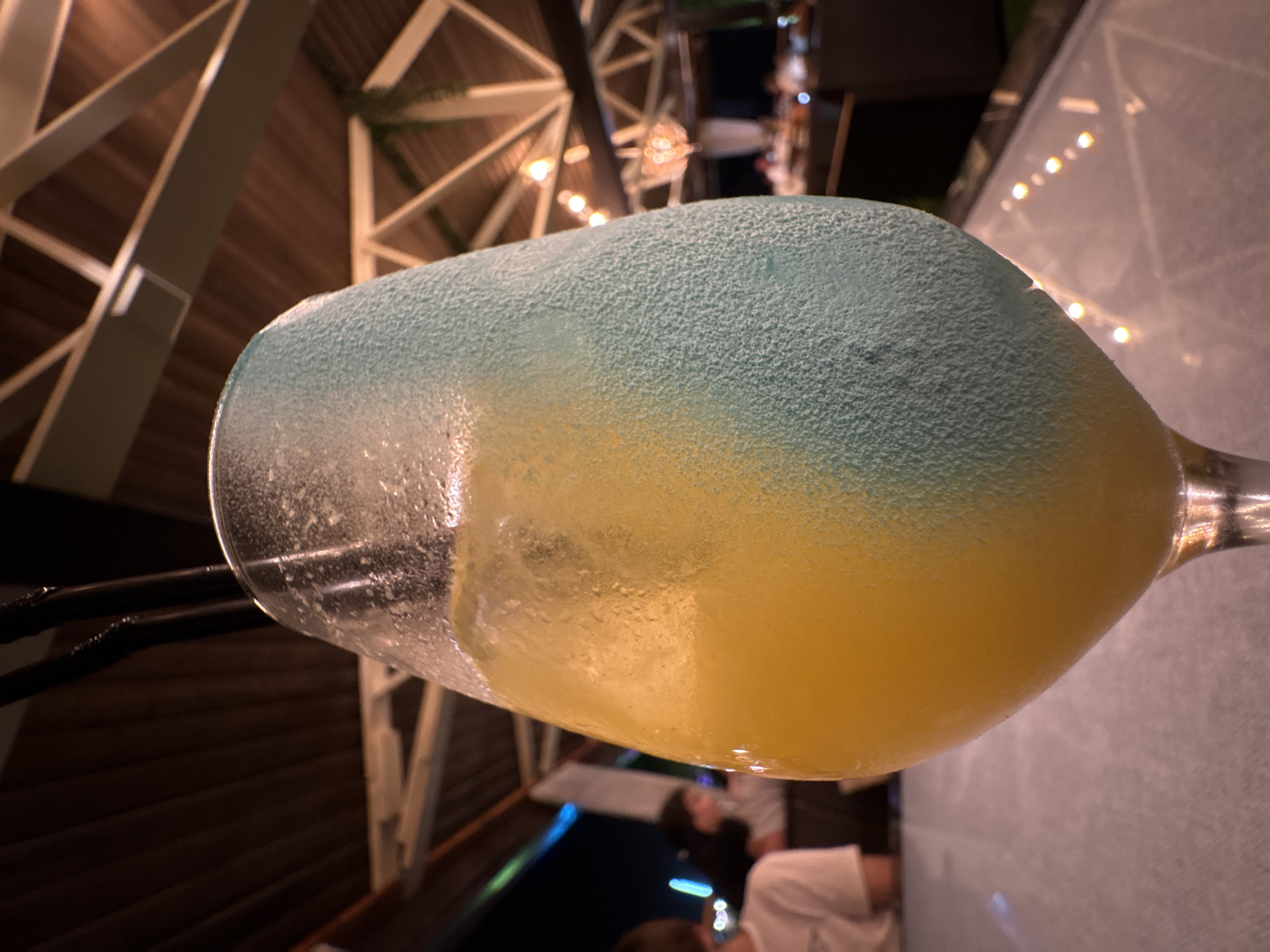 A tall, frosted glass filled with a layered yellow and blue beverage sits on a reflective surface. Two black straws are visible inside the glass. The background suggests an outdoor evening setting with a wooden ceiling structure, lights, and blurred figures of people seated at tables. The scene appears to be at a restaurant or bar. The image is likely taken in Novorossiysk, Russia, given the specified location.