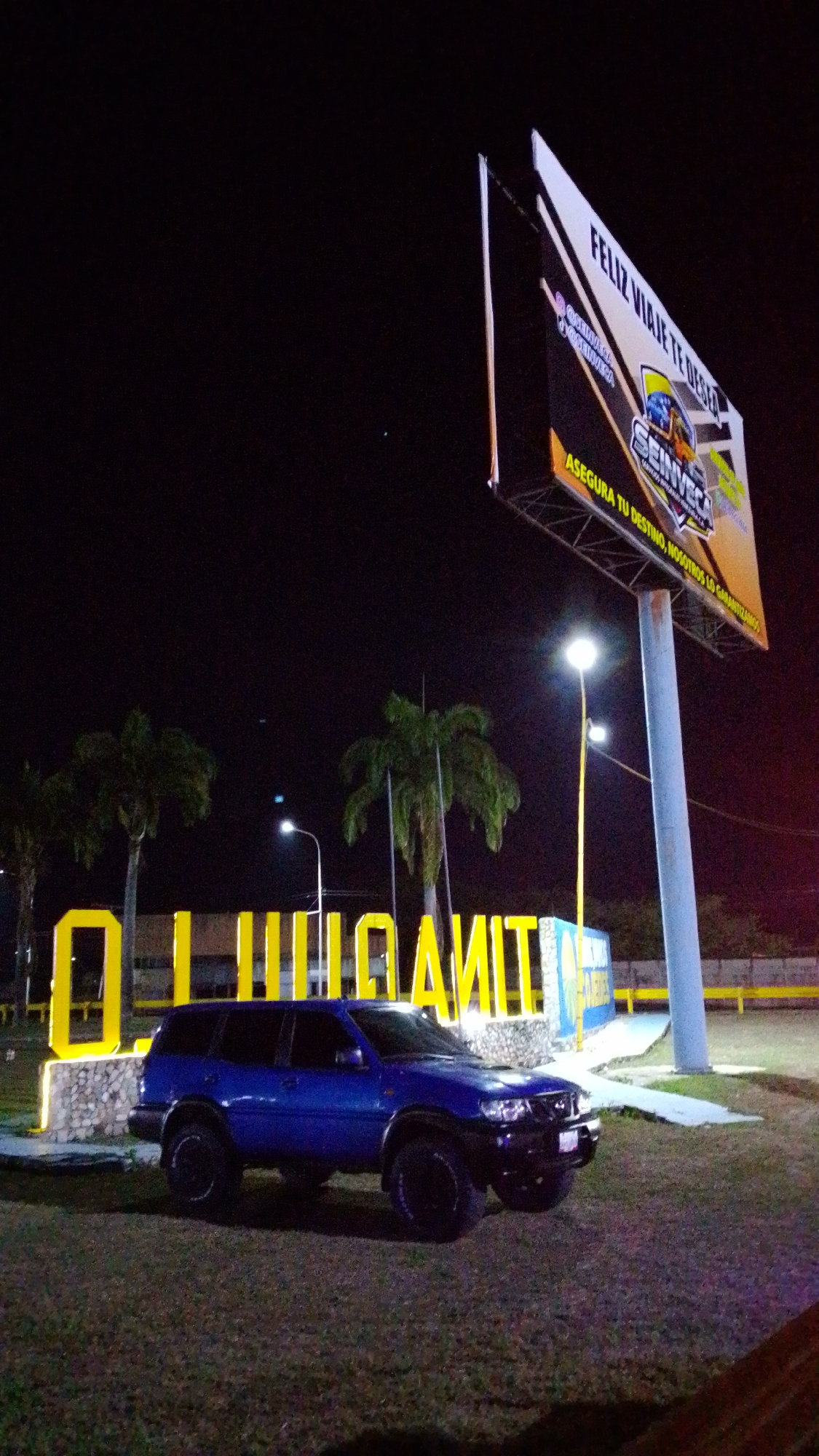 At night in Tinaquillo, Venezuela, a blue Nissan SUV is parked on a grassy area in front of the town's name sign "Tinaquillo" displayed in large yellow block letters. Behind the sign, there are three palm trees and a tall billboard with the Spanish message "FELIZ VIAJE TE DESEA SEINVECA ASEGURA TU DESTINO, NOSOTROS LO GARANTIZAMOS". Two street lights illuminate the scene. A fence is visible in the distance.