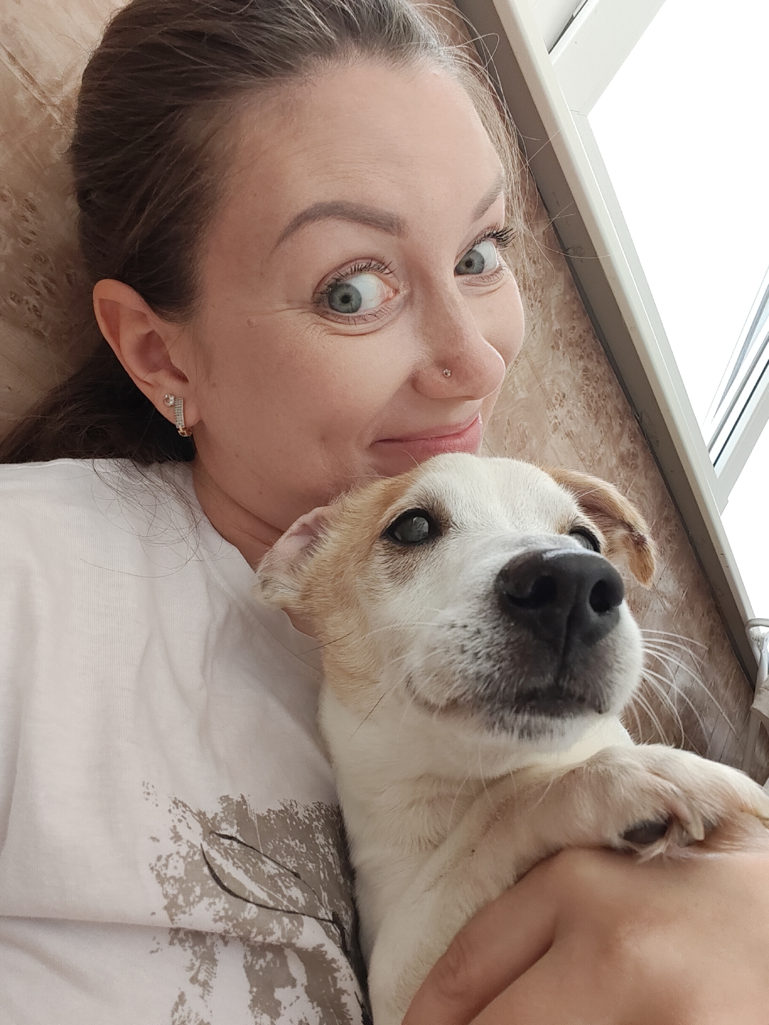 A woman and a small dog are captured in a close-up, indoor shot, likely a selfie. The woman, positioned on the left, has brown hair with some gray strands visible, wide, light-colored eyes (blue/green), and a playful or surprised expression with a slight smile. She wears a small nose stud and small hoop earrings with rhinestones. Her upper body is visible, dressed in a white t-shirt featuring a muted gray and brown graphic design that resembles foliage or abstract shapes.
Cuddled close to the woman, on the right, is a small dog. It has primarily white fur with light brown or tan patches on its ears and around its eyes. The dog has dark eyes, a dark nose, and visible white whiskers. Its front paw is gently resting on the woman's arm. The dog appears calm and looks towards the camera with a gentle expression.
The background consists of a light brown or beige textured surface, possibly a wall with a wood grain or patterned wallpaper. To the right, a white window frame is partially visible, indicating a bright, possibly daylight, setting indoors. No text is visible in the image.