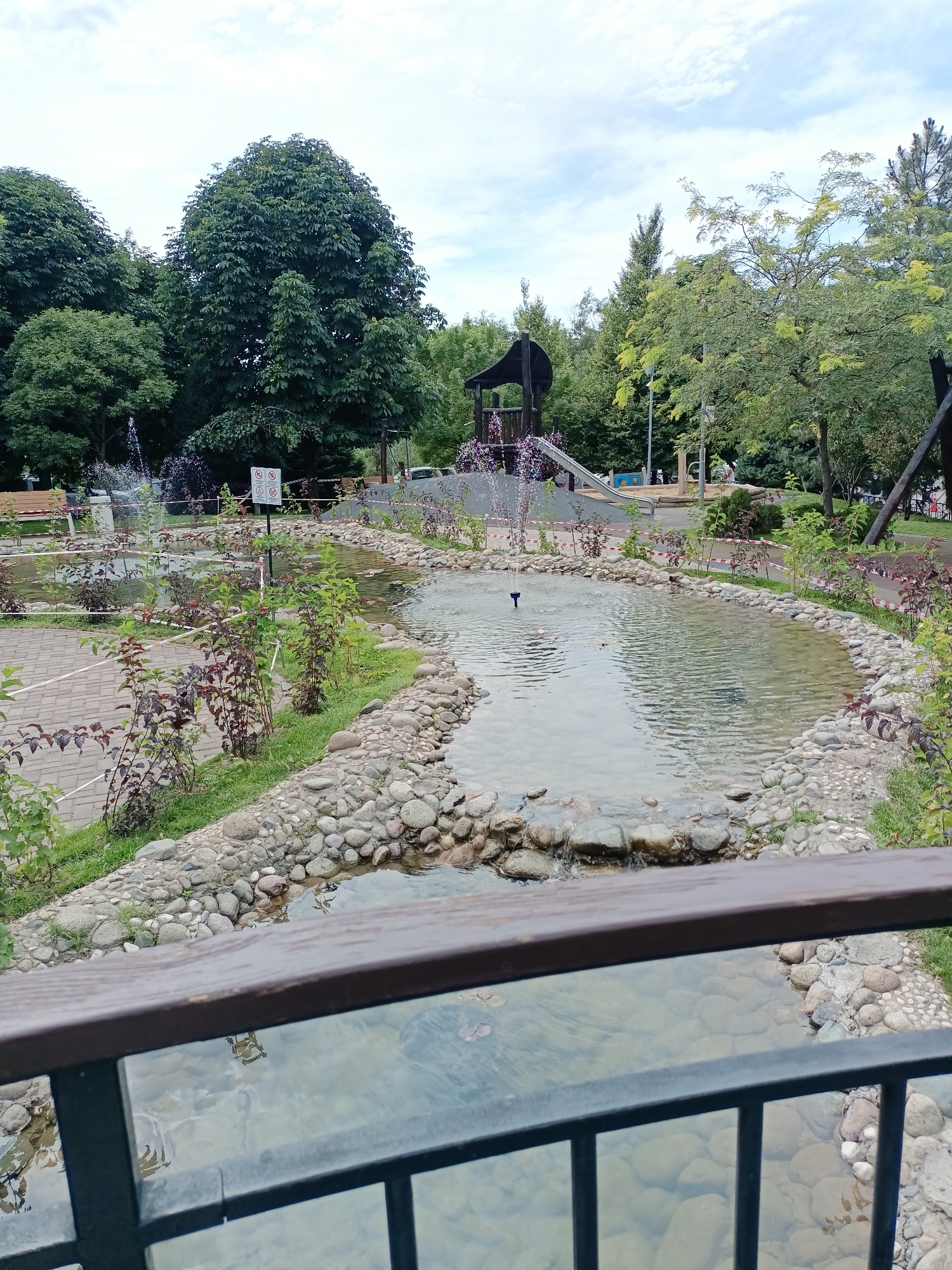 The image captures a serene park scene, likely in Almaty, Kazakhstan, featuring a man-made stream with clear water flowing over a bed of smooth river stones. Small fountains intermittently spray water into the shallow pools, adding a dynamic element to the otherwise tranquil environment. Lush green trees form a dense canopy in the background, providing shade and a natural backdrop. The park includes a children's playground with a slide and other recreational equipment, suggesting it's a family-friendly destination. A paved pathway winds through the park, bordered by neatly planted shrubs with dark purple foliage. A wooden railing in the foreground frames the view, implying the presence of a bridge or elevated walkway. The sky is overcast, with a few scattered clouds, suggesting a pleasant, temperate day.