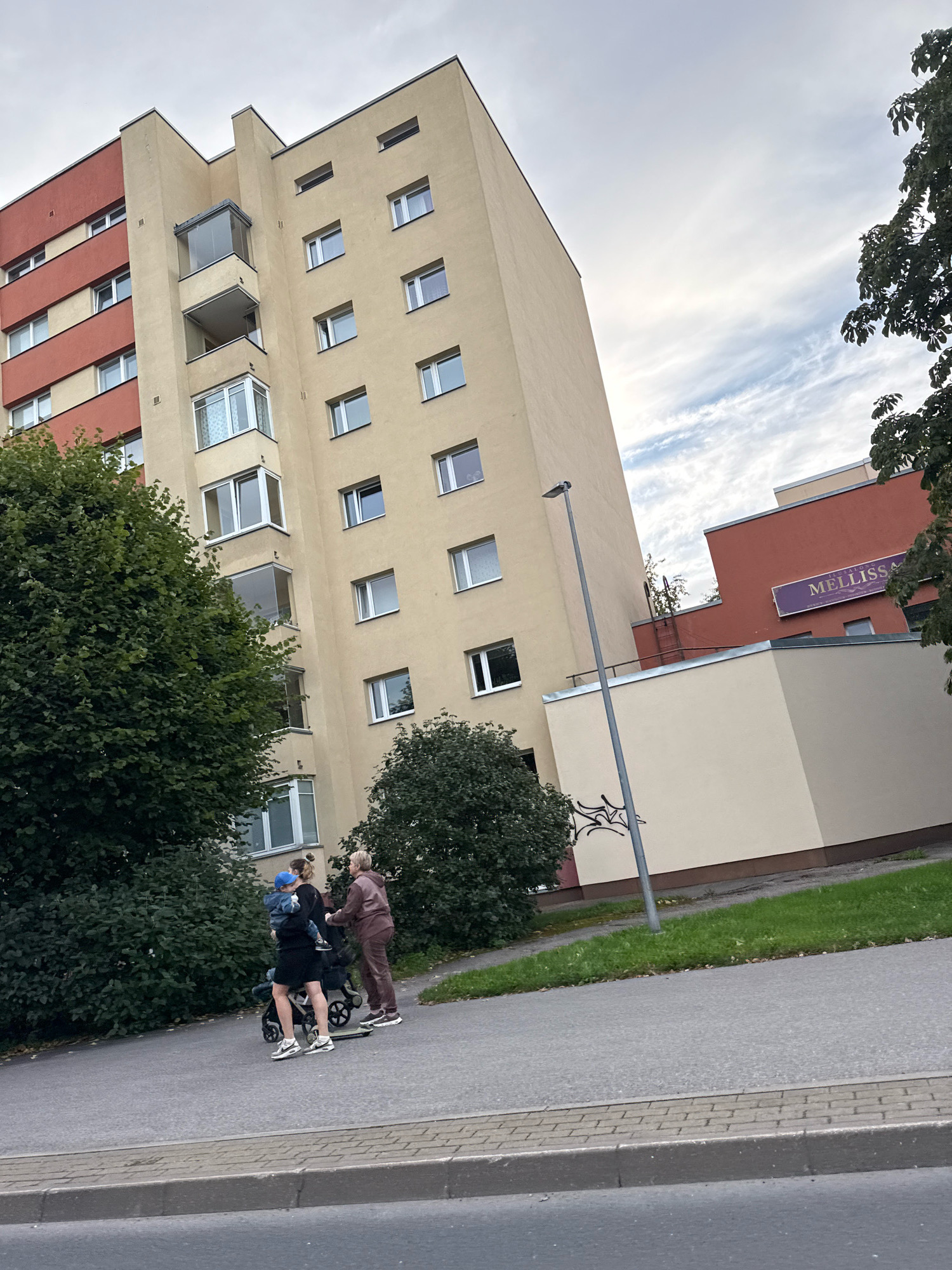 In Tallinn, Estonia, on a cloudy day, a woman and an older woman are walking along a sidewalk next to a large residential building. The woman in the foreground is carrying a young child, who is wearing a blue hat and a denim jacket. She is also pushing a stroller. The older woman, wearing a tracksuit, is walking beside them. The residential building is tall and cream-colored with red accents. To the right of the residential building, a smaller red building has a sign that reads "MĒLLISSĀ". Trees and grass are visible in the background. The sidewalk is paved with grey asphalt and a brick border.
