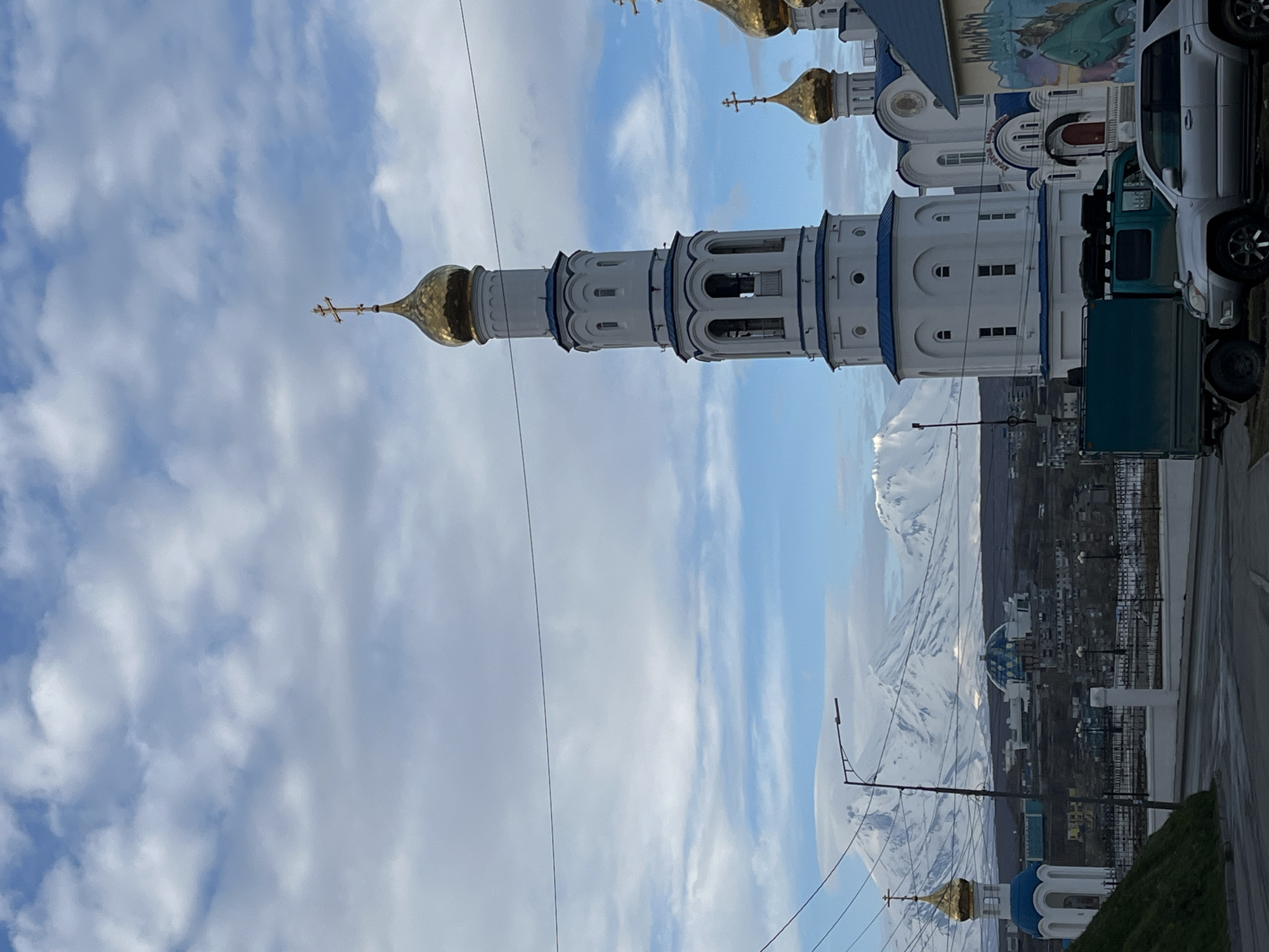 The image depicts a tall, white and blue-trimmed bell tower topped with a golden dome and cross, standing prominently against a sky filled with white and blue clouds. The bell tower is associated with a church, part of which is visible to the right, obscured by parked vehicles. In the background, a city landscape stretches towards snow-capped mountains. This scene is located in Petropavlovsk-Kamchatsky, Russia.