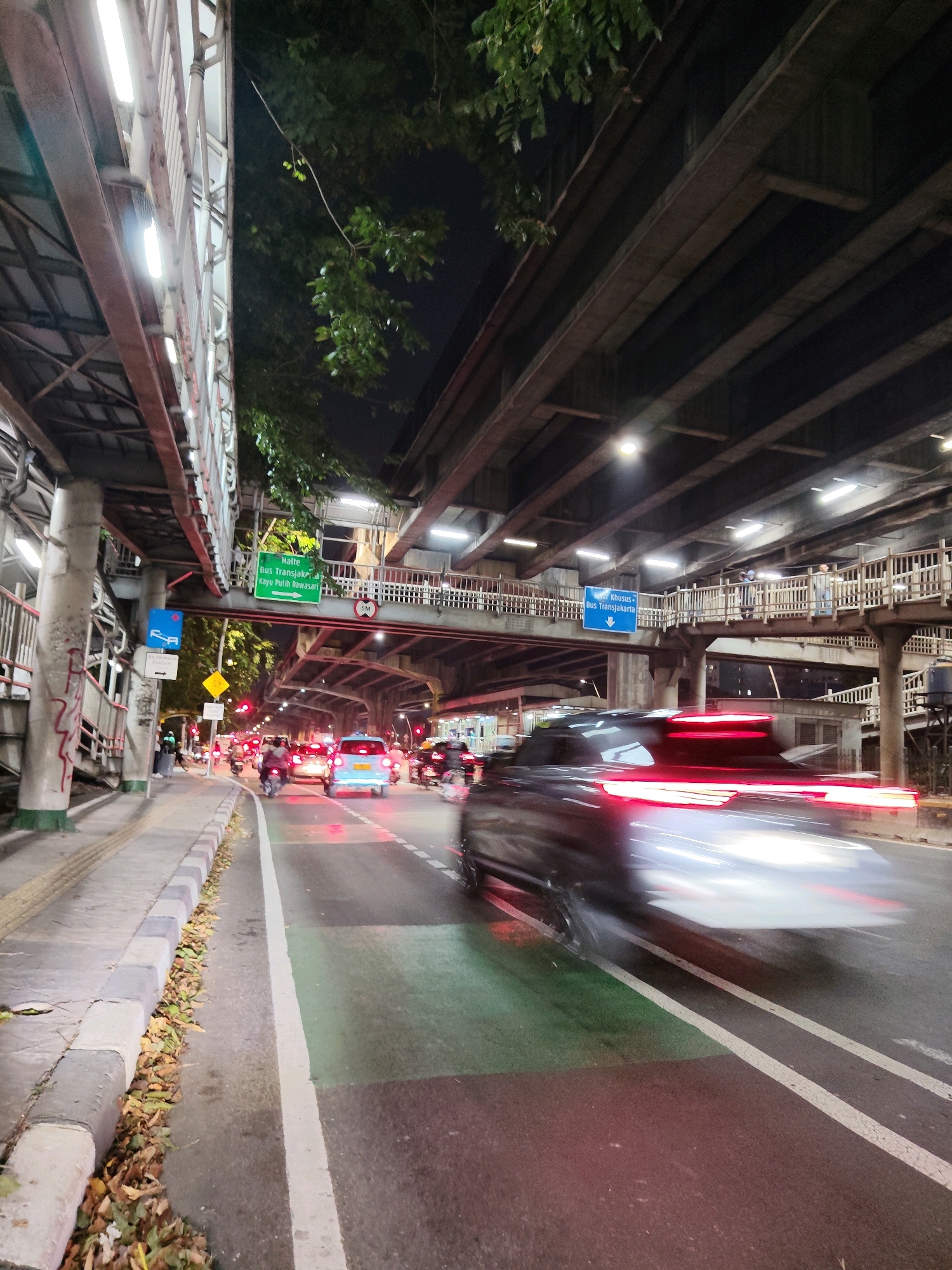 The image depicts an elevated walkway and road scene at night in East Jakarta, Indonesia. The scene is characterized by a busy road with multiple vehicles, including cars and motorcycles, in motion. The street is illuminated by streetlights and the headlights and taillights of vehicles, creating light trails due to motion blur.
On the left side of the road, there is a sidewalk with a concrete barrier and a pedestrian walkway overhead, supported by concrete pillars. The elevated walkway is lined with railings and illuminated by fluorescent lights. Green signage indicating "Halte Bus Transjakarta" (Transjakarta Bus Stop) with an arrow pointing to the right is visible on the elevated structure.
On the right side of the road, another elevated structure, likely part of a highway or overpass, casts shadows on the road below. This structure is also supported by concrete pillars and has lighting fixtures. Pedestrians can be seen on the elevated walkway.
The road itself has painted lane markings, including a green lane, suggesting it might be a dedicated bus lane or a special use lane. Fallen leaves are scattered along the edge of the sidewalk. The overall impression is one of an urban environment at night, with active traffic and pedestrian infrastructure.