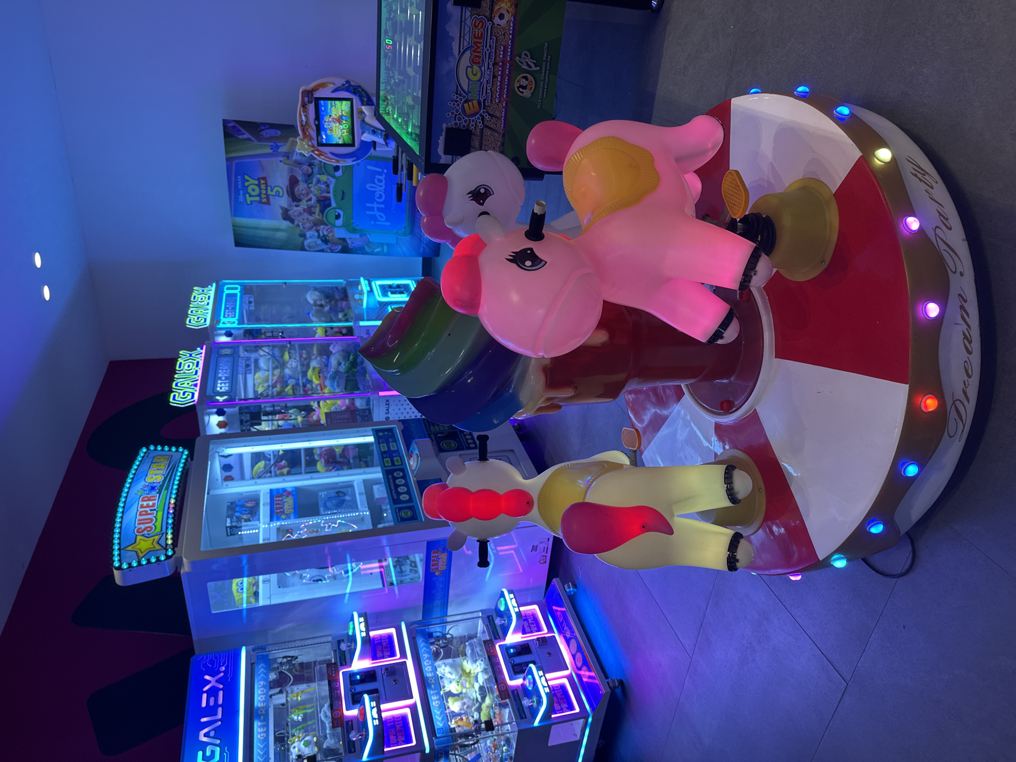 This image captures an indoor entertainment center, likely an arcade or children's play area, illuminated by vibrant artificial lighting. In the foreground, a colorful children's carousel ride dominates the scene. It features three stylized animal figures, two predominantly pink and one yellow and white, resembling ponies or unicorns, each with bright details and black eyes. The carousel's circular base is striped in red and white, encircled by multi-colored LED lights, and subtly inscribed with "Dream Party."
Behind the carousel, an array of arcade game machines and claw machines line the wall, emitting strong blue, purple, and pink neon glows that reflect on the polished floor. A large poster for "Disney/Pixar TOY STORY 5" is prominently displayed, alongside a smaller screen that reads "¡Hola!". One of the arcade machines identifies itself as "EMiGAMES" and lists contact information: "TELS 526-46306, 55649003, 55642700 www.emigames.com.mx". Several machines also bear the brand name "GALEX" and phrases like "<<GET-READY>>", "SUPERSTAR", and "PREMIO".
No people are visible, but the setup suggests a lively environment designed for recreation, particularly for children. The bright, playful aesthetic and Spanish text like "¡Hola!" and "PREMIO" are consistent with entertainment venues in Heroica Puebla de Zaragoza, Mexico. The artificial lighting gives the scene a dynamic, possibly evening, ambiance.