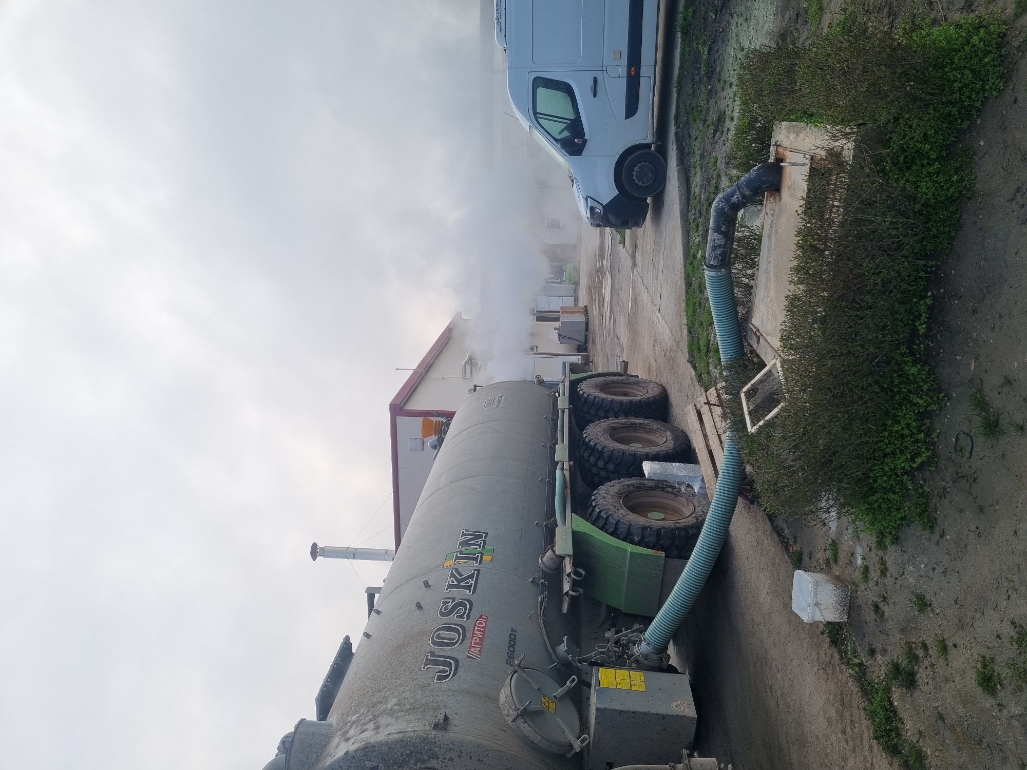 The image shows a gray JOSKIN Agrirton 26000T manure tanker parked on a concrete surface next to a building with a red roof. A white van is parked nearby. A large, blue corrugated hose connects the tanker to a concrete structure near the edge of the concrete. There is a large amount of steam or vapor in the background. The location is suggested to be Ovcharovo, Bulgaria. The ground is dirt. The sky is overcast. Vegetation is growing around the concrete structure.