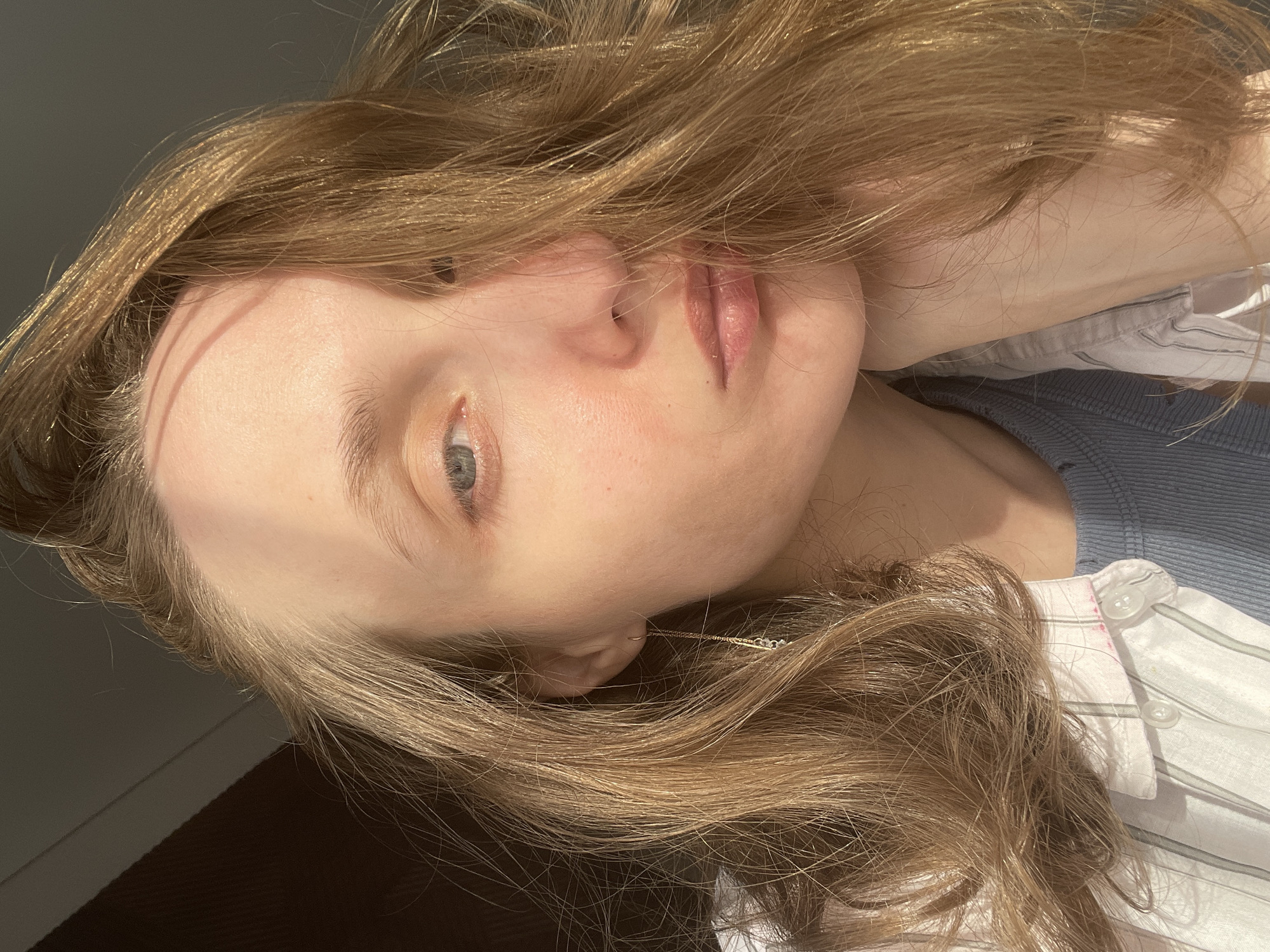 A close-up, slightly upward-angled portrait shows a young woman with light blonde hair, likely indoors. Her hair is long and wavy, partially obscuring her face and shoulders. The woman has pale skin, light blue eyes, and subtle features. She is wearing a light blue t-shirt under a white button-down shirt with thin grey stripes. Part of her right hand is visible near her neck. The background is a dark brown, possibly a wall or furniture. The lighting is bright, casting strong shadows on her face and hair. There's no information in the image to indicate that the picture was taken in Sepych, Russia.