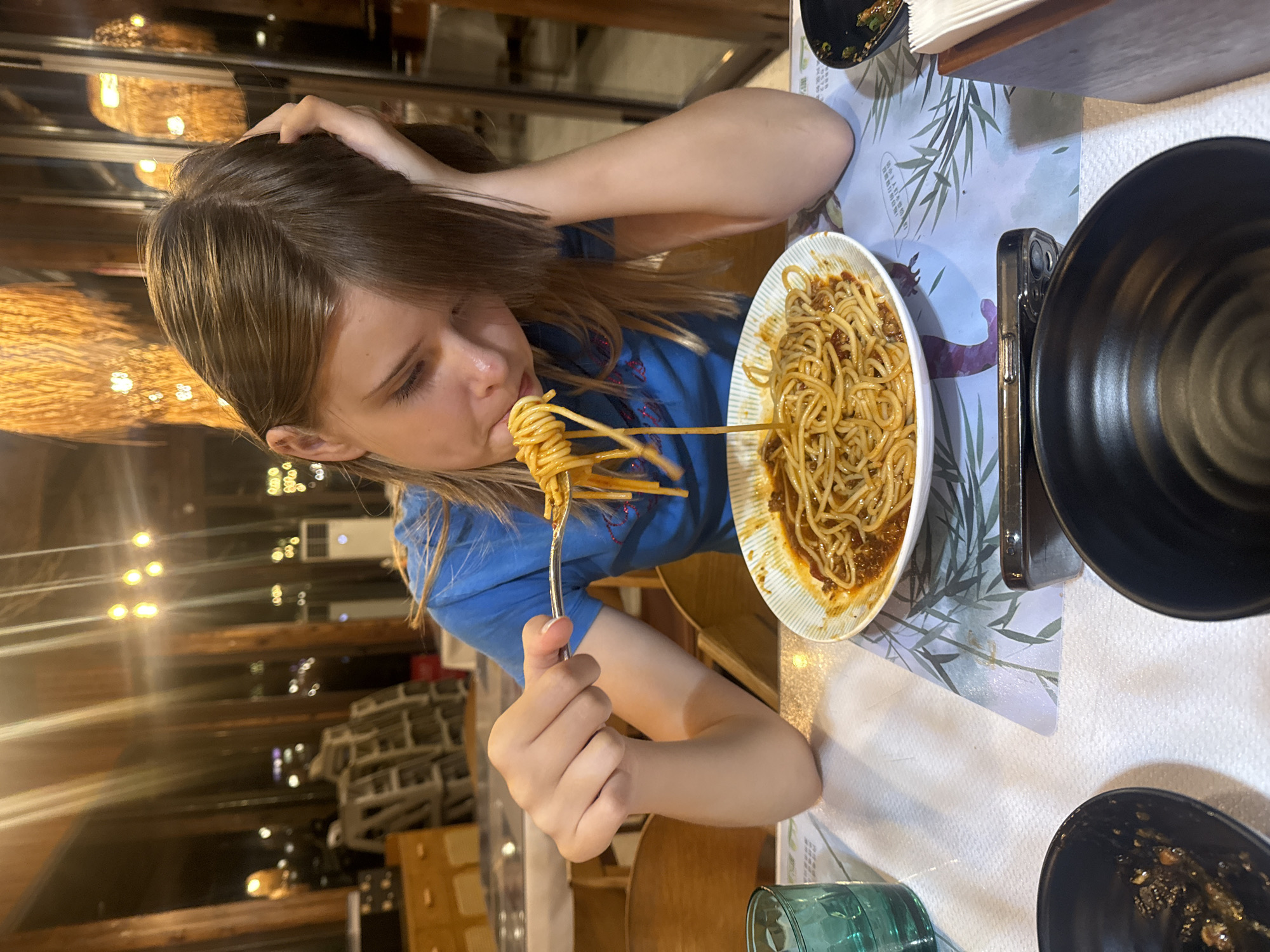 A young person with shoulder-length brown hair is seated at a table, eating spaghetti from a white and blue striped bowl. They are wearing a blue t-shirt and have their left hand on their head. The spaghetti is being lifted to their mouth with a fork. The table is covered with a white tablecloth and a placemat with a bamboo pattern. There are black bowls on the table, one of which appears to be partially empty. A green glass is to the left of the bowl of spaghetti. The background shows a dining area with wooden beams and warm lighting. The location is Wuxing District, China.