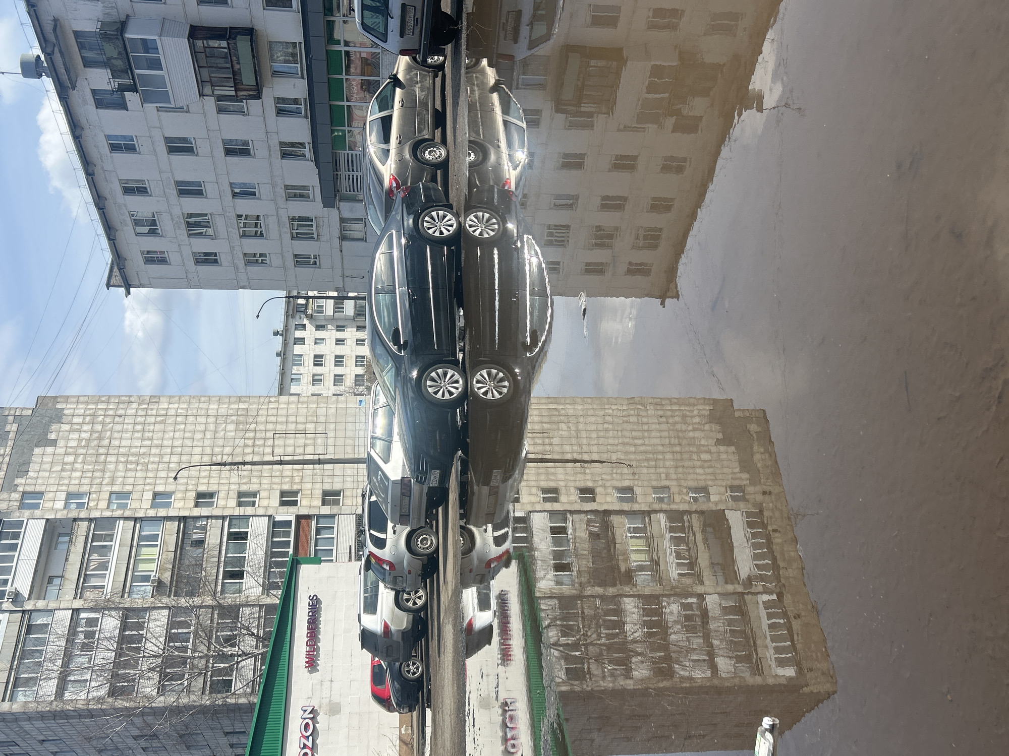 The image shows a street scene reflected in a large puddle in the city of Chaikovsky, Russia. Several parked cars are lined up along the street, including a red car, a white car, a gray car, and a black car. The reflection also shows the facades of two multi-story apartment buildings. One building has rows of windows and a sign on the ground floor reading "OZON" and "WILDBERRIES". The puddle's surface reflects the sky, with a few clouds visible. The image is oriented in a way that the objects and buildings on the street are vertical, while their reflection is oriented horizontally.