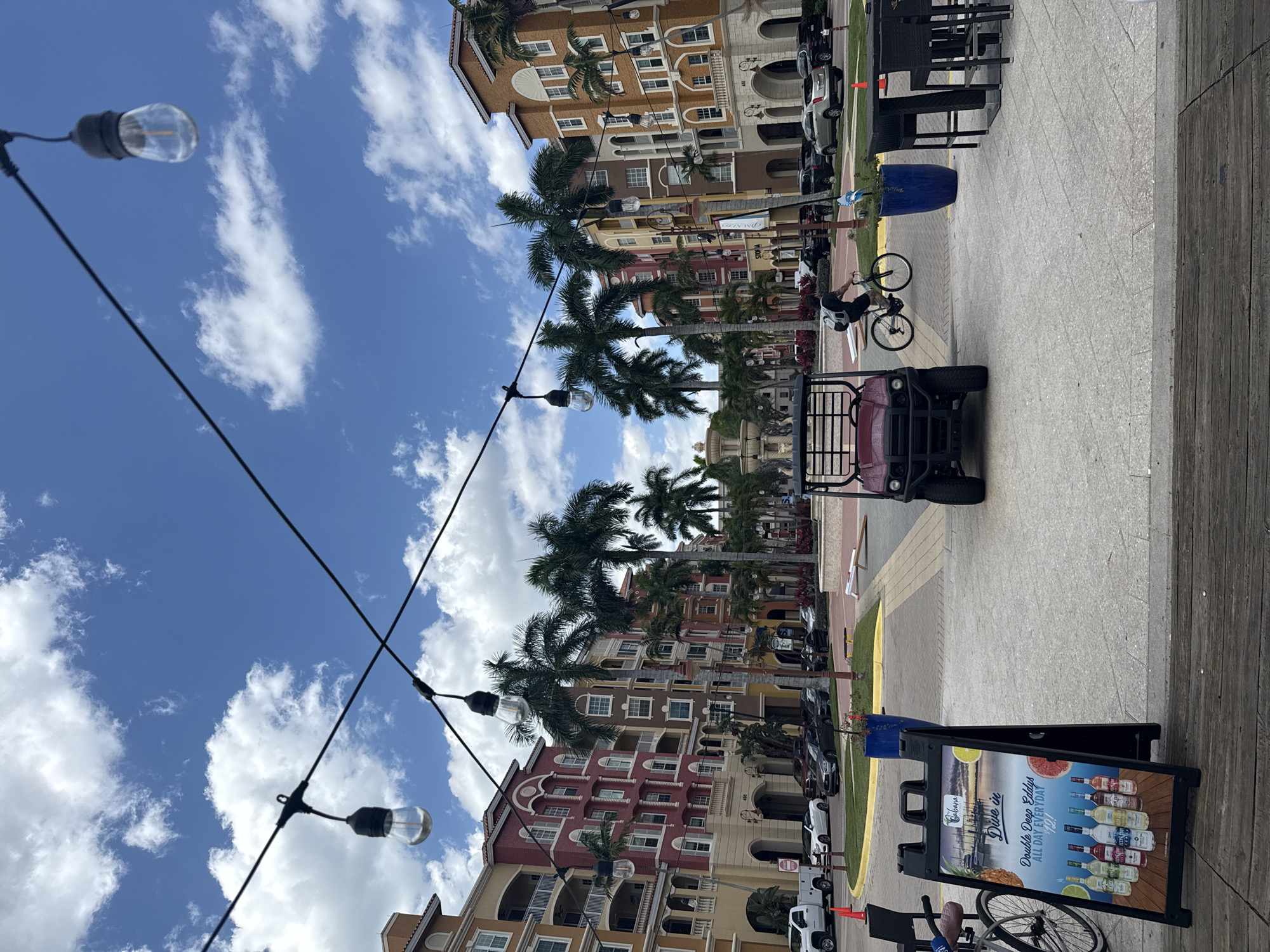 The image depicts a street scene in Naples, Florida, USA, under a partly cloudy sky. Multi-story buildings with diverse architectural styles and colors line one side of the street, interspersed with palm trees. Parked cars are visible along the building side of the street. Overhead, string lights with exposed bulbs are strung across the street.
On the right side of the frame, near the bottom, is a sidewalk with a wooden plank edge. A sandwich board sign advertising alcoholic beverages is placed on the sidewalk. Further along the sidewalk, there is a red utility vehicle with an open back and a bicycle ridden by a person with a backpack. There are tables and chairs visible in the background. A large blue planter sits by the side walk.