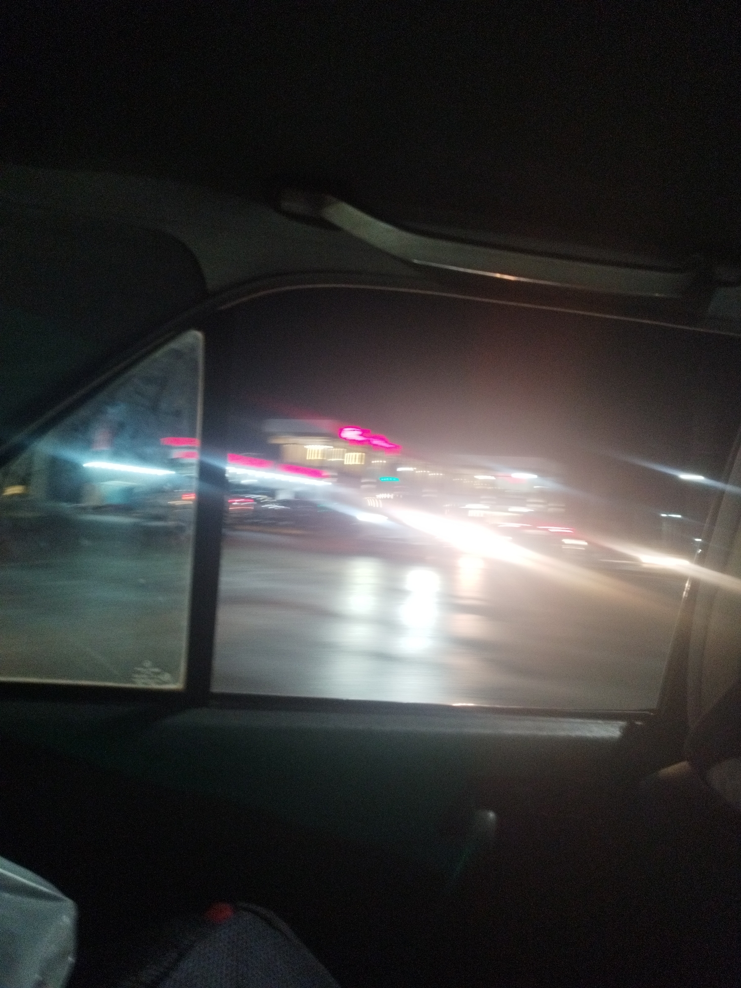 The image captures a blurred night-time view from inside a vehicle in Shymkent, Kazakhstan. Through the car window, a brightly lit commercial building with pink neon signage is visible. The street appears wet, reflecting the lights from the building and passing vehicles, suggesting it may have recently rained. The motion blur indicates the car is in transit. The interior of the car is dark, with only a small portion of the dashboard and a seat visible.