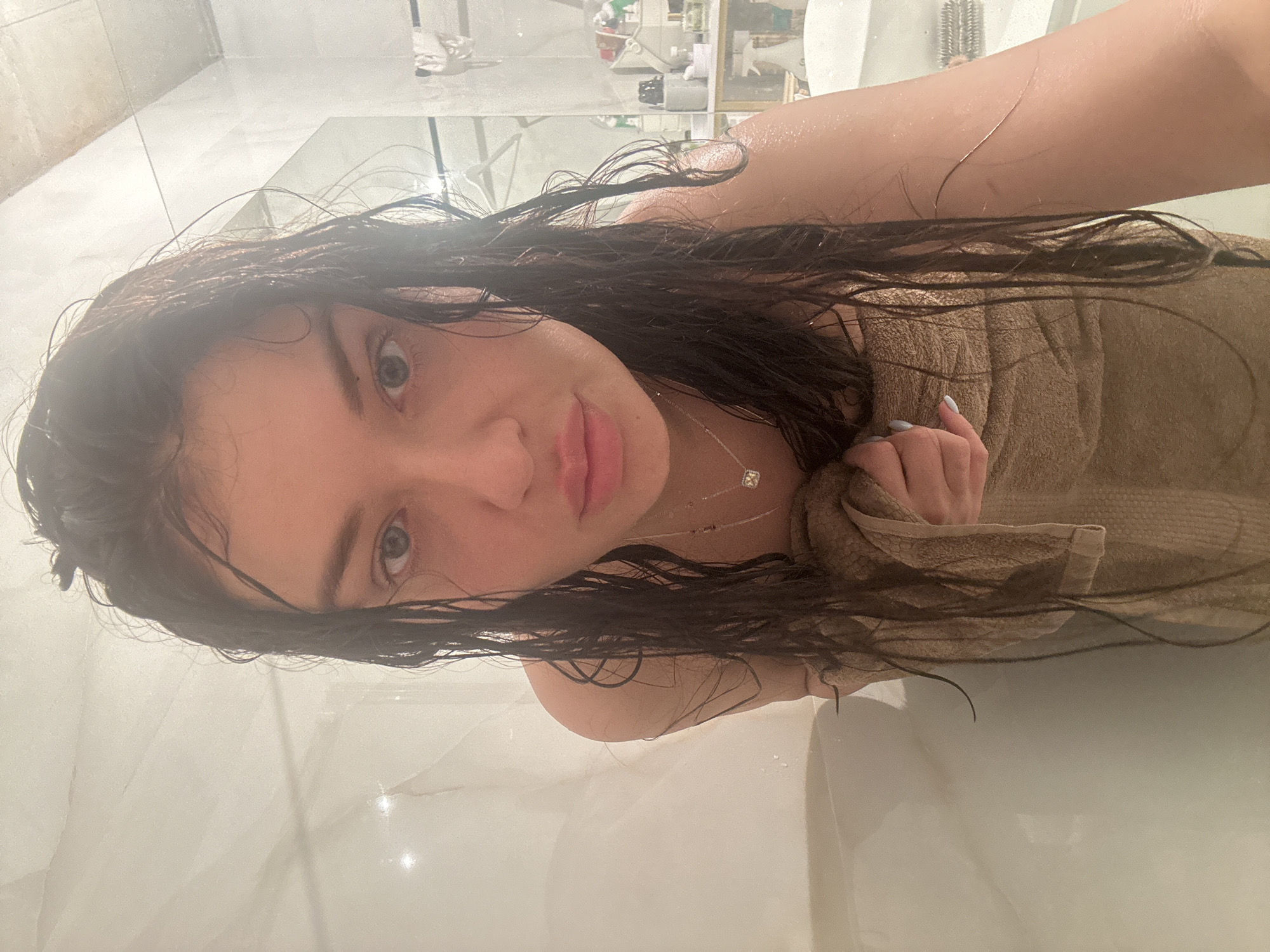The image displays a young woman taking a selfie in what appears to be a modern bathroom, set in Moscow, Russia. The photo is oriented vertically, with the woman's head to the left and her body extending to the right. Her dark, wet hair is draped around her face and shoulder, indicating she has recently showered or bathed, a detail reinforced by water droplets visible on her skin.
She has light-colored eyes and full lips, looking directly at the camera with a calm, slightly smiling expression. A delicate silver necklace with a square-cut pendant adorns her neck. She is wrapped in a light brown or beige towel, which she holds with one hand, revealing light-colored painted fingernails.
The background features glossy, light-colored tiled walls and surfaces, possibly marble, giving the bathroom a clean and bright aesthetic. Reflections in the shiny surfaces show glimpses of other bathroom items, such as bottles and a brush, further confirming the setting. The lower portion of the image shows a white bathtub or shower basin. The lighting is bright and artificial, contributing to the clear and pristine appearance of the scene. No visible text is present in the image.