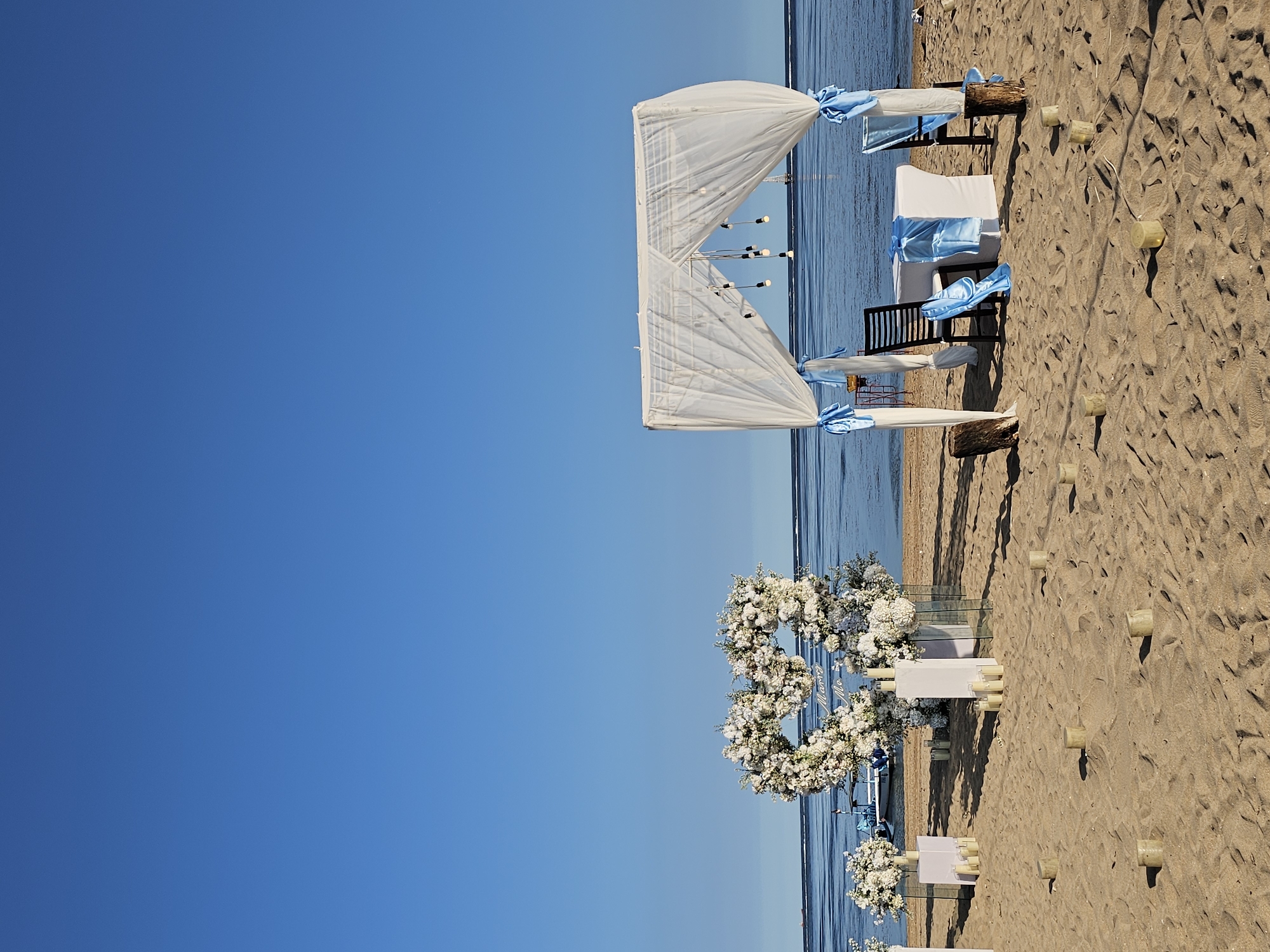 The image captures a beach wedding setup in Denpasar, Indonesia. The scene features a white canopy draped with light blue ribbons, positioned near the shoreline. Two chairs are placed underneath the canopy, adorned with similar ribbons. On the sand, floral arrangements in the shape of infinity symbols and flower boxes are set up. Numerous white candles are scattered on the sand along a pathway leading toward the canopy. The environment includes a clear blue sky and the ocean in the background. No people are visible in the scene.