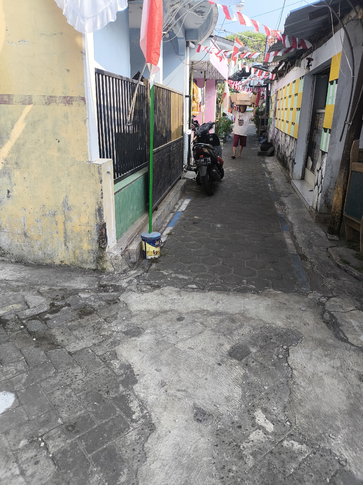 The image depicts a narrow alleyway, likely in Lumajang, Indonesia, adorned with red and white Indonesian flags strung across it, suggesting a national celebration or event. The alley is paved with interlocking hexagonal concrete blocks and bordered by buildings on both sides. On the left, a yellow-painted wall with weathered textures and a black metal fence with a green pole and a blue bucket at its base is visible. A motorcycle is parked on the left side of the alley. Further down the alley, a person is walking away from the camera, carrying boxes stacked on their head and wearing a white t-shirt and red shorts. The buildings lining the alley show various architectural styles and paint colors, with some featuring decorative patterns and others appearing more utilitarian. The overall scene conveys a sense of everyday life in a densely populated urban environment, enhanced by the festive decorations.