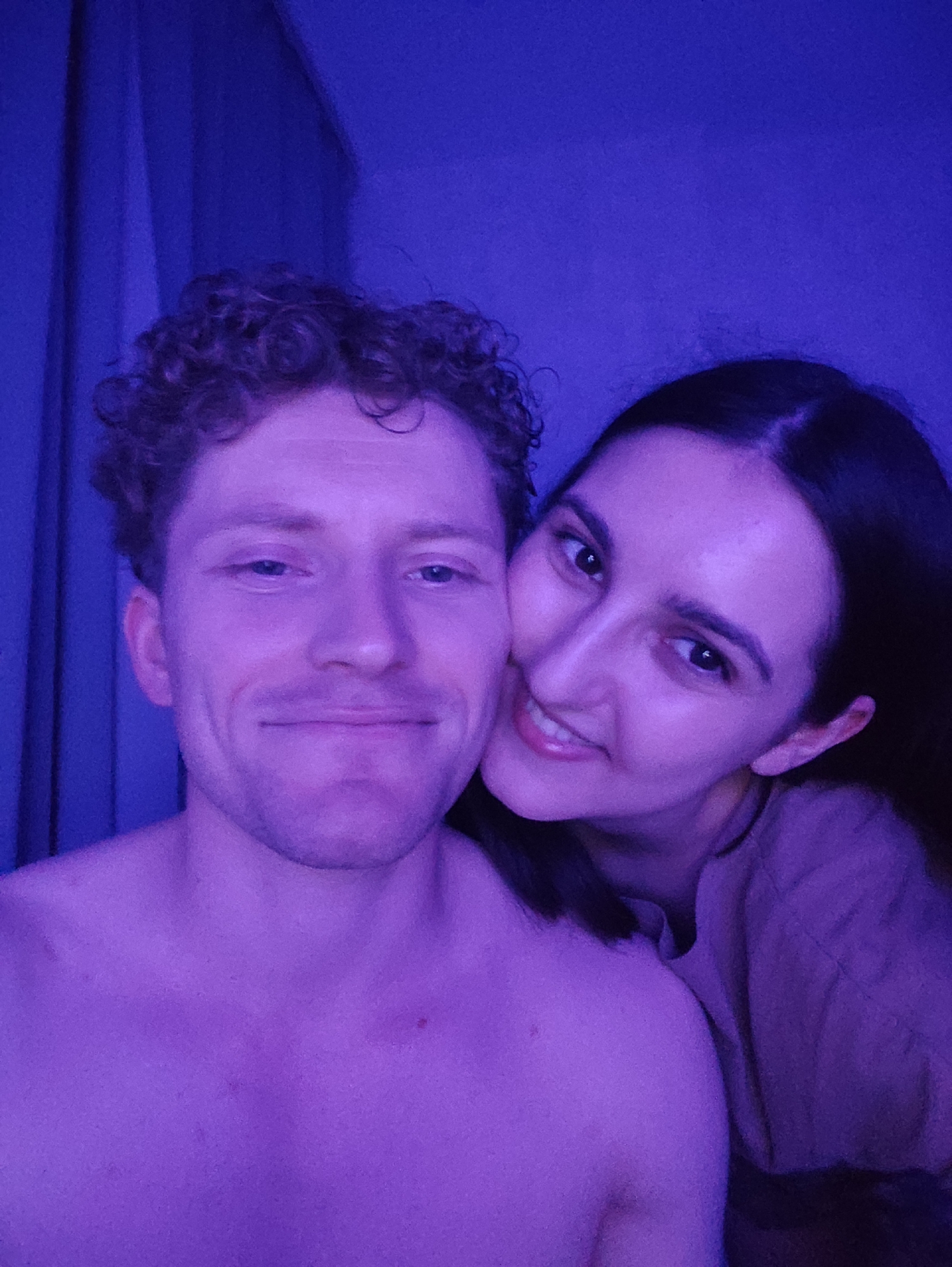 In an indoor setting illuminated by purple lighting, a close-up selfie captures a man and a woman. The man, in the foreground and on the left, has curly light brown hair and is shirtless, smiling. To his right and slightly behind him, the woman has dark brown hair and is smiling and looking at the camera. She is wearing a light brown garment. The background consists of a dark curtain on the left and a dimly lit wall on the right. The overall ambiance suggests an intimate or casual moment.