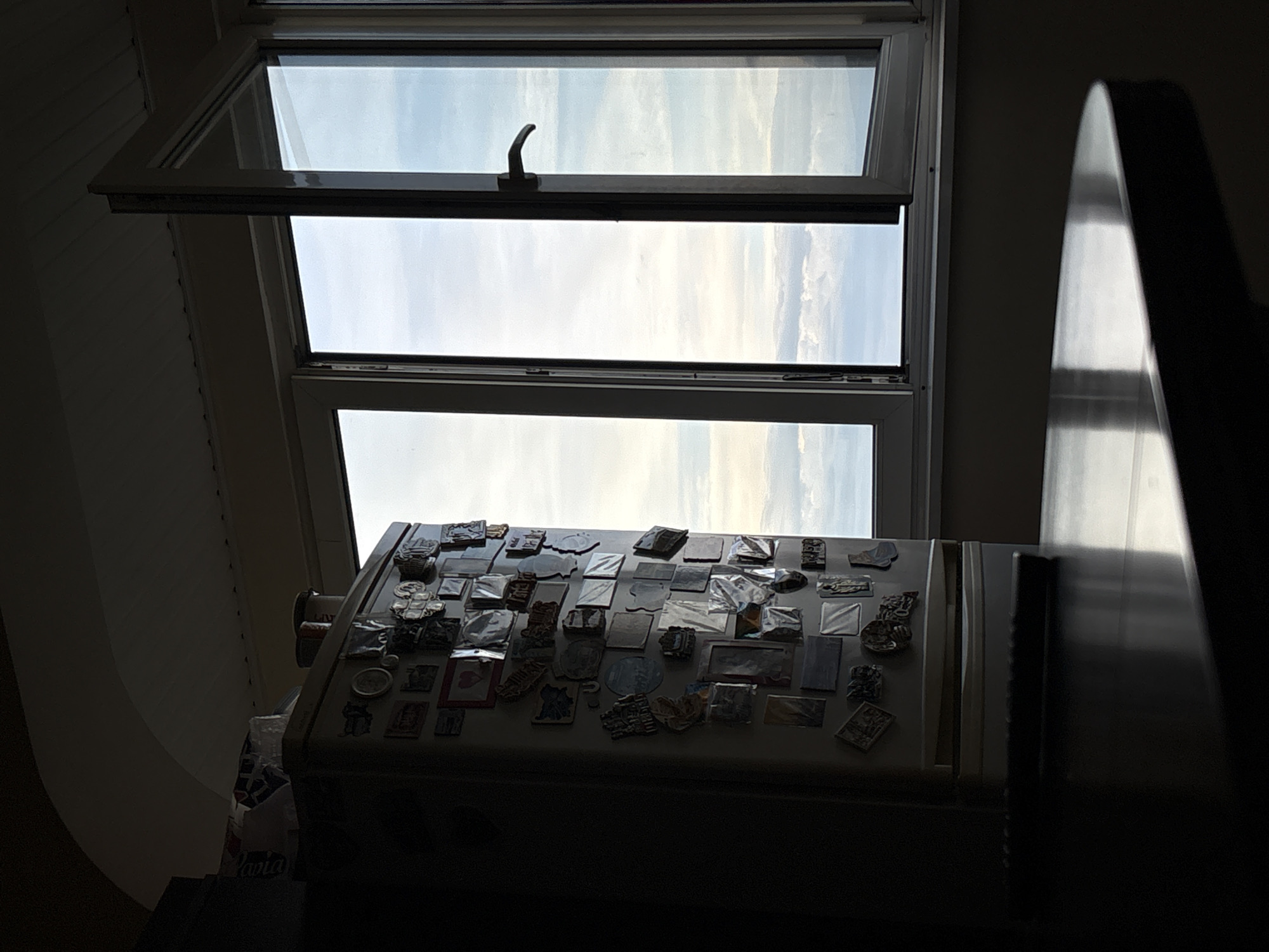 The image depicts an interior scene with a refrigerator covered in various magnets, positioned next to a large window. The window is open, revealing a cloudy sky with a hint of blue. A dark, reflective surface, likely a table, occupies the foreground. The lighting suggests it is daytime, with the sun casting long shadows. The setting appears to be a room in Vratsa, Bulgaria. There are no people visible in the image. The refrigerator is white and stands to the left of the window. The window frame is white, and the glass panes are visible. The overall atmosphere is quiet and domestic.