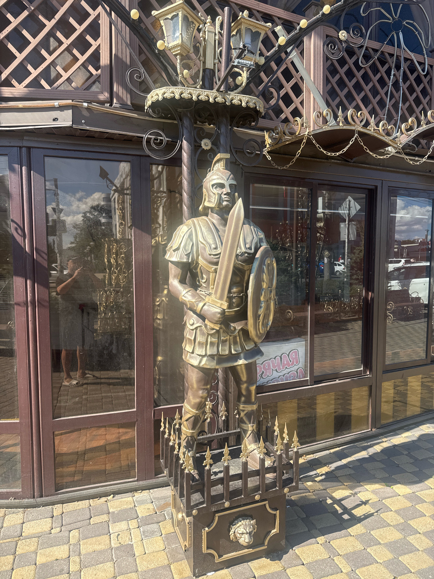 The image shows a bronze statue of a Roman soldier in front of a building with large glass windows. The soldier is holding a sword and shield and is wearing armor. The building appears to be a storefront or restaurant, with ornate metalwork above the windows. In the reflection of the glass, a person can be seen taking a photo of the statue. The ground is paved with interlocking stones. The statue is positioned on a decorative metal pedestal with a lion's head motif. The scene is illuminated by bright sunlight, casting distinct shadows on the ground. The sign visible through the window reads "Прямая линия," which translates to "Hotline" or "Direct line" in Russian, suggesting a service-oriented establishment. The context provided indicates this location is in Anapa, Russia.