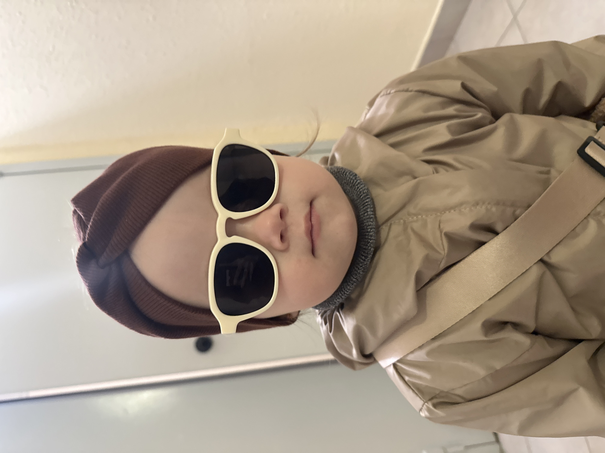 The image features a close-up portrait of a young child, likely a toddler, looking directly at the camera with a stylish and confident demeanor.
**Subjects:** The central subject is a child.
**Objects:** The child is wearing oversized, light-cream or beige-framed sunglasses with dark lenses. They are dressed in a dark brown knit beanie or hat and a puffy, beige or light tan winter coat. A grey, ribbed neck warmer or turtleneck is visible around their neck. A wide, beige fabric strap, potentially from a bag or part of the coat, crosses the child's torso on the right side of the frame.
**Scene/Setting:** The background is simple and indistinct, suggesting an indoor environment with plain, light-colored walls. A subtle horizontal line in the background hints at an architectural feature like a door frame or skirting. The image is captured from a slightly low angle.
**Activity/Event:** The child is posing for a photograph, maintaining a direct gaze at the viewer (through their sunglasses). There is no specific activity taking place.
**Notable Details:**
* **Emotions:** The child's expression is neutral, conveying a sense of coolness or cuteness, amplified by the large sunglasses.
* **Time of day/Weather:** The warm attire—coat, hat, and neck warmer—suggests the photo was taken during cooler weather, likely autumn or winter. The time of day is not discernible from the indoor setting.
* **Location cues:** There are no specific visual cues within the image to identify the location. However, external context indicates the photo was taken in Yekaterinburg, Russia.
* **Visible text:** No text is visible in the image.
The original media file is oriented in landscape, rotated 90 degrees counter-clockwise. When viewed upright, the child's head is slightly tilted, and they are prominently framed.