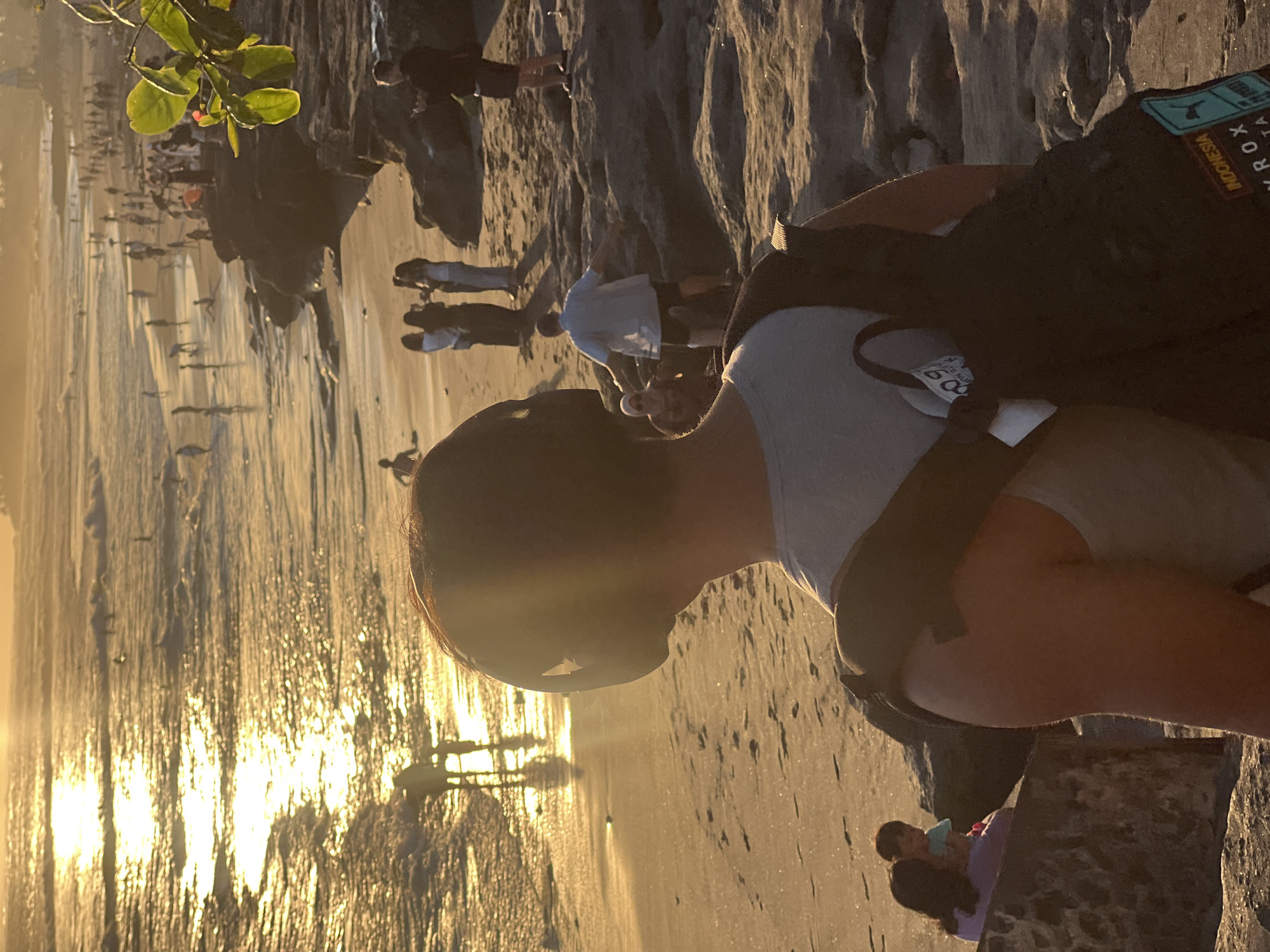 An outdoor, medium shot captures a beach scene at sunset in North Kuta, Indonesia. In the foreground, a person is facing away from the camera, wearing headphones and a gray shirt, with a backpack slung over their shoulders. Behind this person, the beach is populated with many individuals, some walking along the shoreline and others wading in the ocean. The sun, low in the sky, casts a warm golden glow on the water, creating a shimmering reflection. Rocky outcrops line the edge of the beach, and a few people are visible standing or sitting on these rocks. The overall atmosphere is one of leisure and enjoyment of the coastal environment at dusk.
