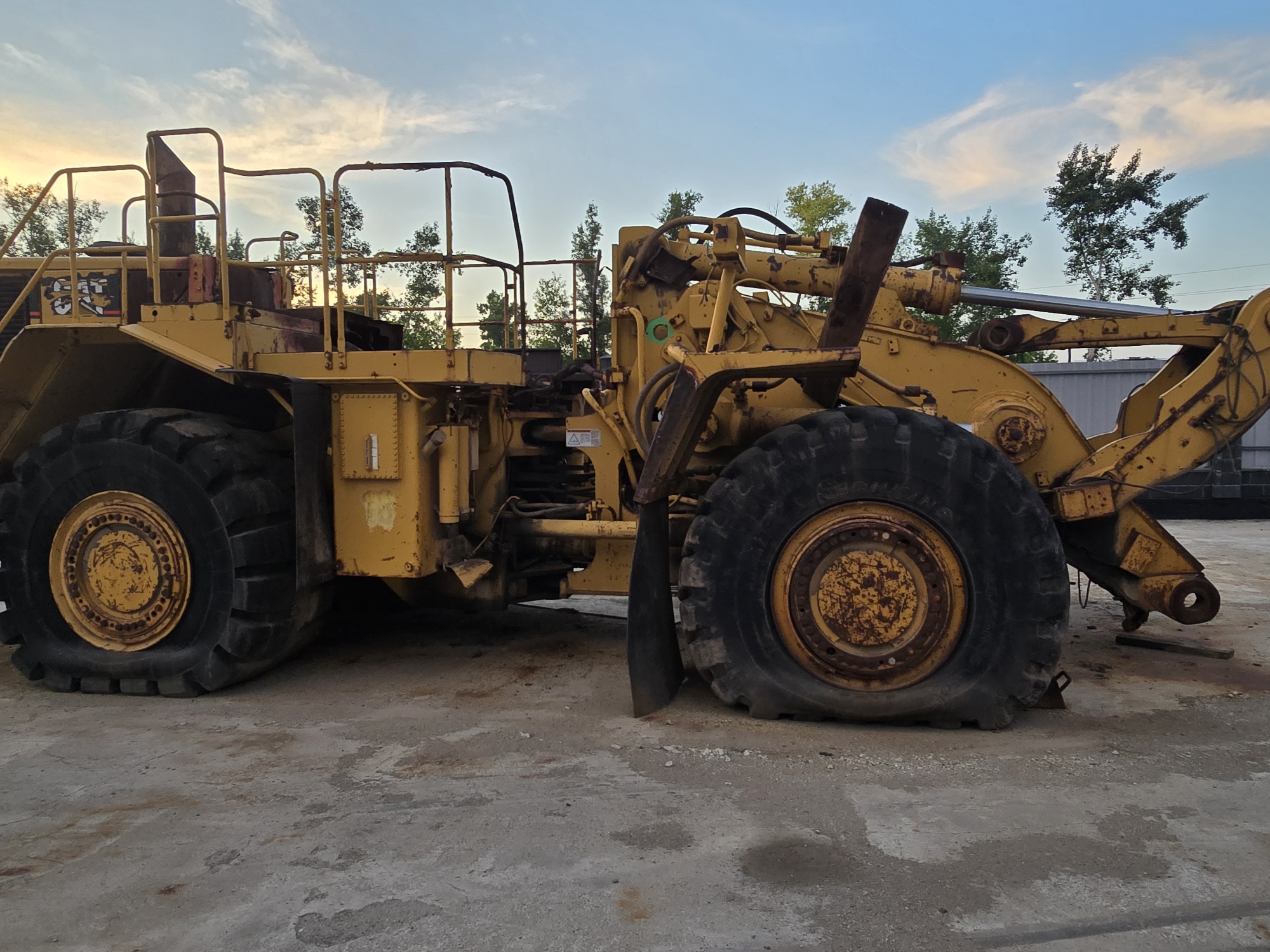 The image shows a large, yellow, industrial vehicle, possibly a front-end loader or a mining truck, parked outdoors on a concrete surface. The vehicle appears to be old and weathered, with visible signs of rust and wear on its tires and body. The sky is partly cloudy with hints of sunset colors. Trees are visible in the background. The vehicle is in a state of disrepair; its tires are flat and appear to be deflated. The scene does not suggest any specific activity or interaction, but rather depicts a stationary, possibly abandoned, piece of heavy machinery. There are no people visible in the image. The location is identified as Oskemen, Kazakhstan, suggesting an industrial or construction site.