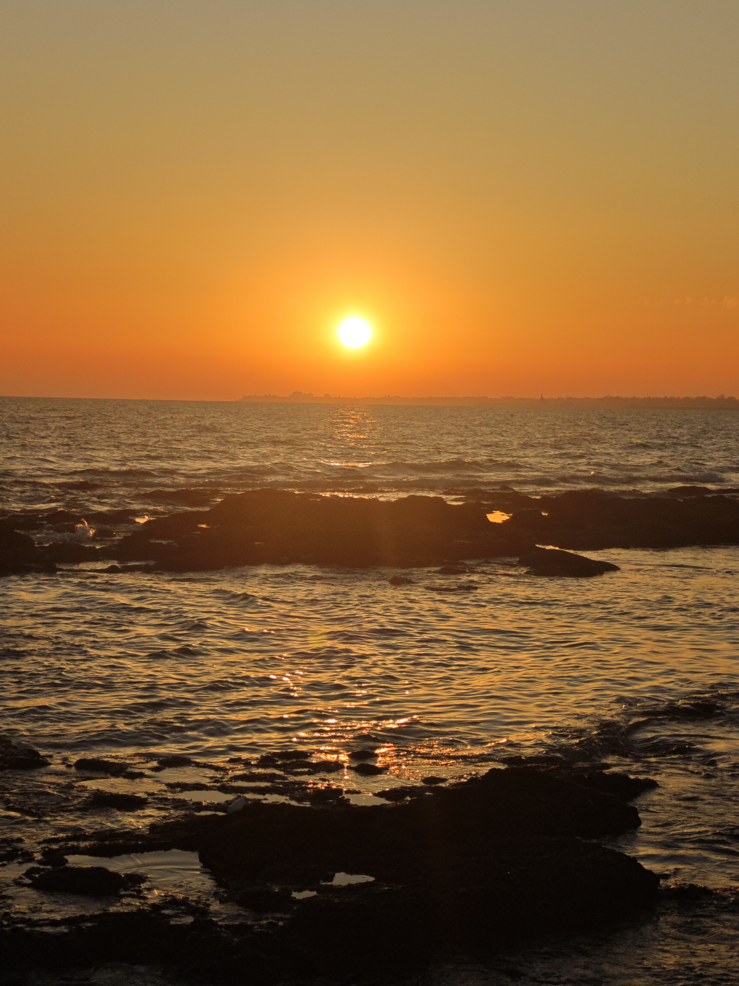 A vertical, low-angle shot depicts a sunset over the sea in Costa Oeste, Spain. The sun, a bright white orb, is positioned high in the orange sky, casting a golden reflection on the rippling water. Dark, rocky formations emerge from the sea in the foreground, with some submerged and others forming small islands. The distant horizon is dotted with faint silhouettes of buildings, suggesting a city or town. The water is a deep blue-grey, with sparkling highlights from the setting sun. The overall mood is tranquil and serene, capturing the beauty of a coastal sunset.