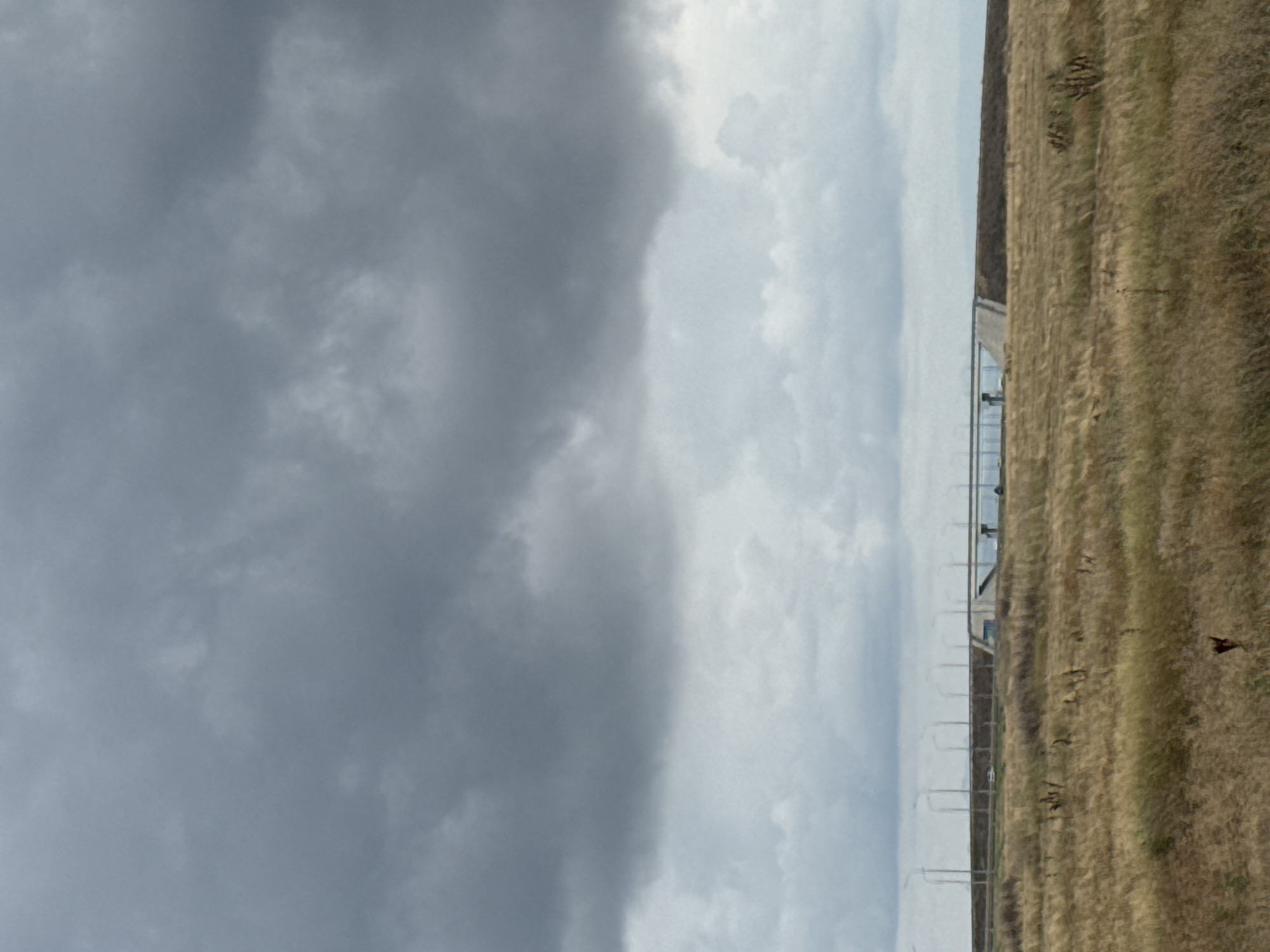 The image depicts a wide-angle view of a barren landscape under a heavy, overcast sky. In the foreground, dry, golden grass covers the ground, interspersed with sparse, dark brown vegetation. In the mid-ground, a concrete overpass stretches horizontally across the scene. The overpass is supported by pillars and features a raised roadway. To the left of the overpass, a line of streetlights is visible, stretching into the distance. A single white car is seen on the roadway of the overpass. The background is dominated by a vast expanse of gray clouds, creating a somber atmosphere. The scene is devoid of human figures. The overall impression is one of desolation and open space, possibly a rural or undeveloped area on the outskirts of a city. The provided location of Батальненское сельское поселение, Ukraine, suggests this is likely a scene from that region.