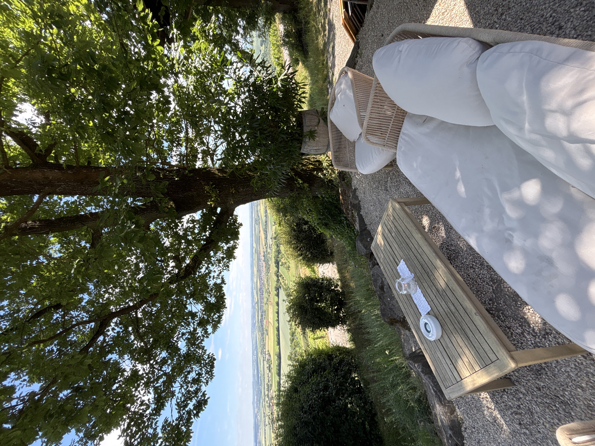 In an outdoor setting with a panoramic view of a rolling landscape under a clear blue sky, a white cushioned outdoor sofa and two rattan chairs with white cushions are arranged on a gravel patio. A wooden table with a glass candle holder, a white patterned runner, and an ashtray sits in front of the sofa. Green bushes and the trunk and canopy of a large tree with green leaves dominate the foreground and middle ground. The landscape beyond consists of fields, vineyards, and distant buildings. The overall impression is one of a tranquil, scenic outdoor seating area.