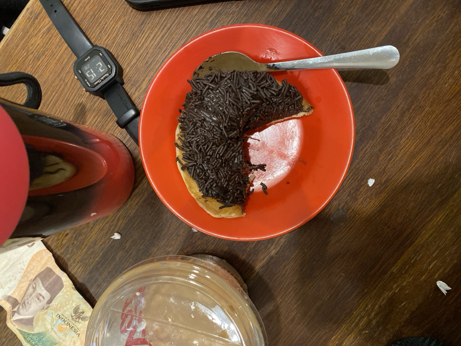 The image presents an overhead view of a dark brown wooden table, presumably in Pontianak, Indonesia. A bright orange circular plate centrally features a partially consumed crescent-shaped pancake, densely covered with dark brown chocolate sprinkles (mese). A silver spoon rests on the right side of the plate, partially embedded in the sprinkles. To the left of the plate, a clear plastic container with a domed lid contains a light brown substance. Behind the plate, a dark metallic or black insulated mug with a reflective surface and a red lid is visible. A crumpled 10,000 Indonesian Rupiah banknote is positioned to the left of the mug. To the right of the mug, a black Alexandre Christie digital wristwatch, displaying "2:15", lies on the table. A portion of a black object, likely a smartphone, is visible at the far right edge of the frame. Small white specks, consistent with food crumbs, are scattered on the tabletop.
