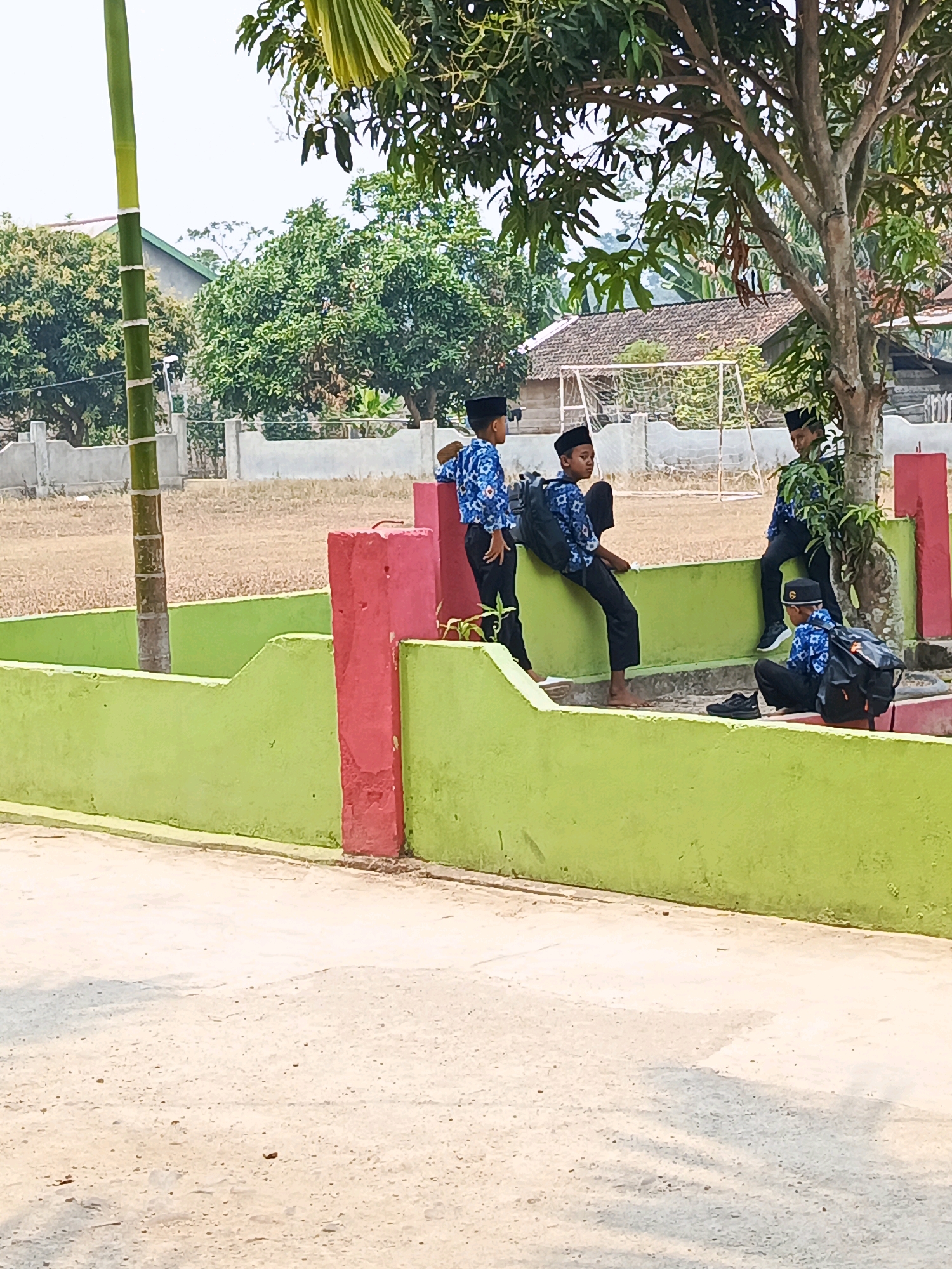 In Megang Sakti V, Indonesia, four young boys, dressed in blue and white patterned shirts, black pants, and black skullcaps, are gathered in an outdoor area. One boy stands near a green and pink wall, while another sits on the wall, looking towards the right. A third boy is seated on the ground to the right of the wall, with a backpack beside him. The fourth boy leans against a tree on the right side of the frame, also with a backpack. The background shows a field with a soccer goal, trees, and houses. A bamboo pole stands to the left of the scene, and the ground is paved with a light-colored, sandy material. The lighting suggests it is daytime, with shadows visible on the ground.
