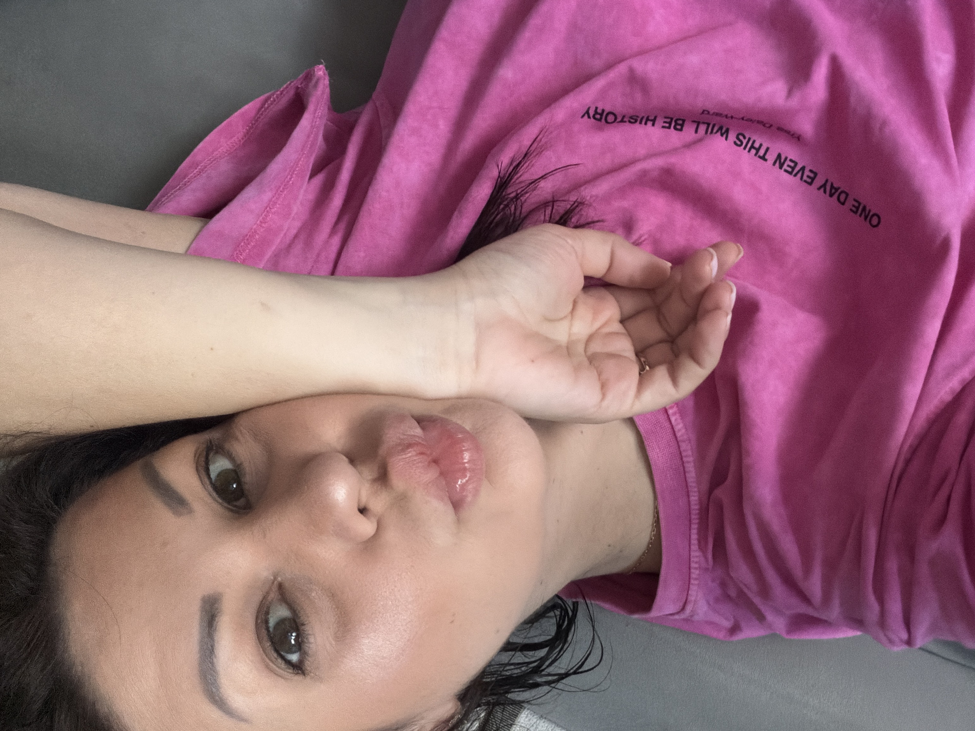 A woman lies on her side on a grey surface, possibly a sofa or bed, her head tilted and her right arm across her face, obscuring part of her cheek and mouth. She is wearing a bright pink t-shirt with a curved black text print on the right side that reads "ONE DAY EVEN THIS WILL BE HISTORY". Her dark hair is wet and rests on her shoulder. She appears to be making a kissing gesture with her lips. Her left eye is open, looking towards the camera, while her right eye is partially obscured by her arm. The lighting suggests an indoor setting. No specific location in Sochi, Russia is discernible from the image.
