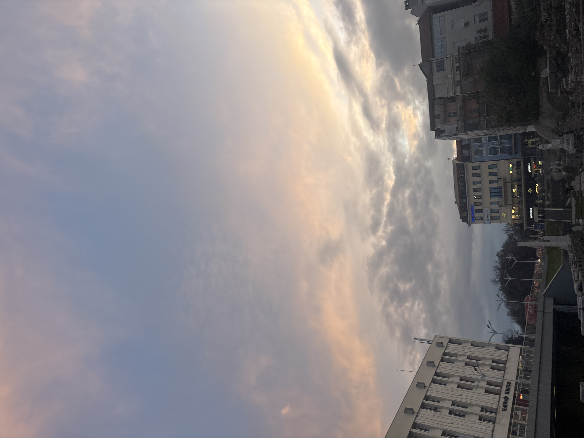 The image depicts a view in Plovdiv, Bulgaria at dusk. The sky occupies most of the frame, with streaks of light orange and grey clouds. Below the sky are a few buildings, including the building of Plovdiv Drama Theater. The buildings are various heights and colors. A part of the Ancient Roman Stadium of Philippopolis is seen between the buildings. Trees with bare branches can be seen lining the streets. The lighting suggests it is either sunrise or sunset.