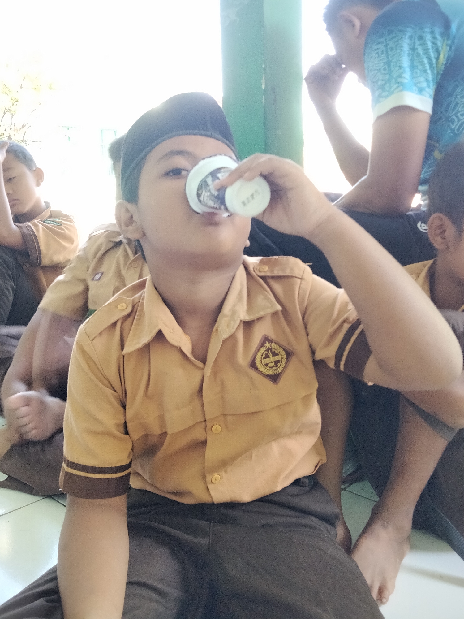 The image shows a group of young boys sitting down, possibly during a break or an outdoor activity. The boy in the foreground, wearing a tan uniform and a black peci (a cap worn by Muslim men and boys), is drinking from a small white cup. He is looking directly at the camera. Another boy to his left is wearing a similar tan uniform and has his hand raised to his head. A third boy is visible in the background, wearing a blue shirt with a white pattern.
The setting appears to be outdoors, with bright light suggesting it is daytime. The background is blurred but hints at greenery and possibly a building structure. The boys are dressed in what seem to be school uniforms, indicated by the tan shirts with insignia and dark pants. The overall mood is casual and relaxed. The white cup in the foreground has some text on it, but it is difficult to decipher clearly from the image. The location is Pucakwangi, Indonesia.