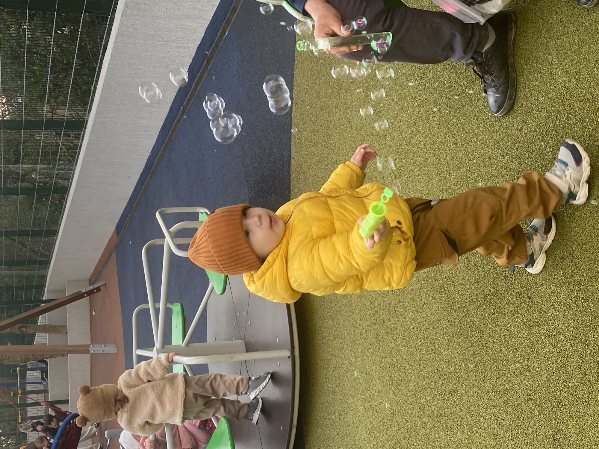 A young child wearing a yellow puffer jacket and a brown hat stands on a green, artificial turf surface with brown pants and sneakers. The child is holding a green bubble wand in their right hand, seemingly in the act of blowing bubbles. Several bubbles are visible in the air around the child. Above the child, an adult wearing dark pants and black shoes is also blowing bubbles with a clear bubble wand. To the left of the child, a carousel-like structure with green seats is present, and another child wearing a bear-eared hat is standing on it. The background includes a blue and gray wall. The location is a playground, potentially in Sochi, Russia.