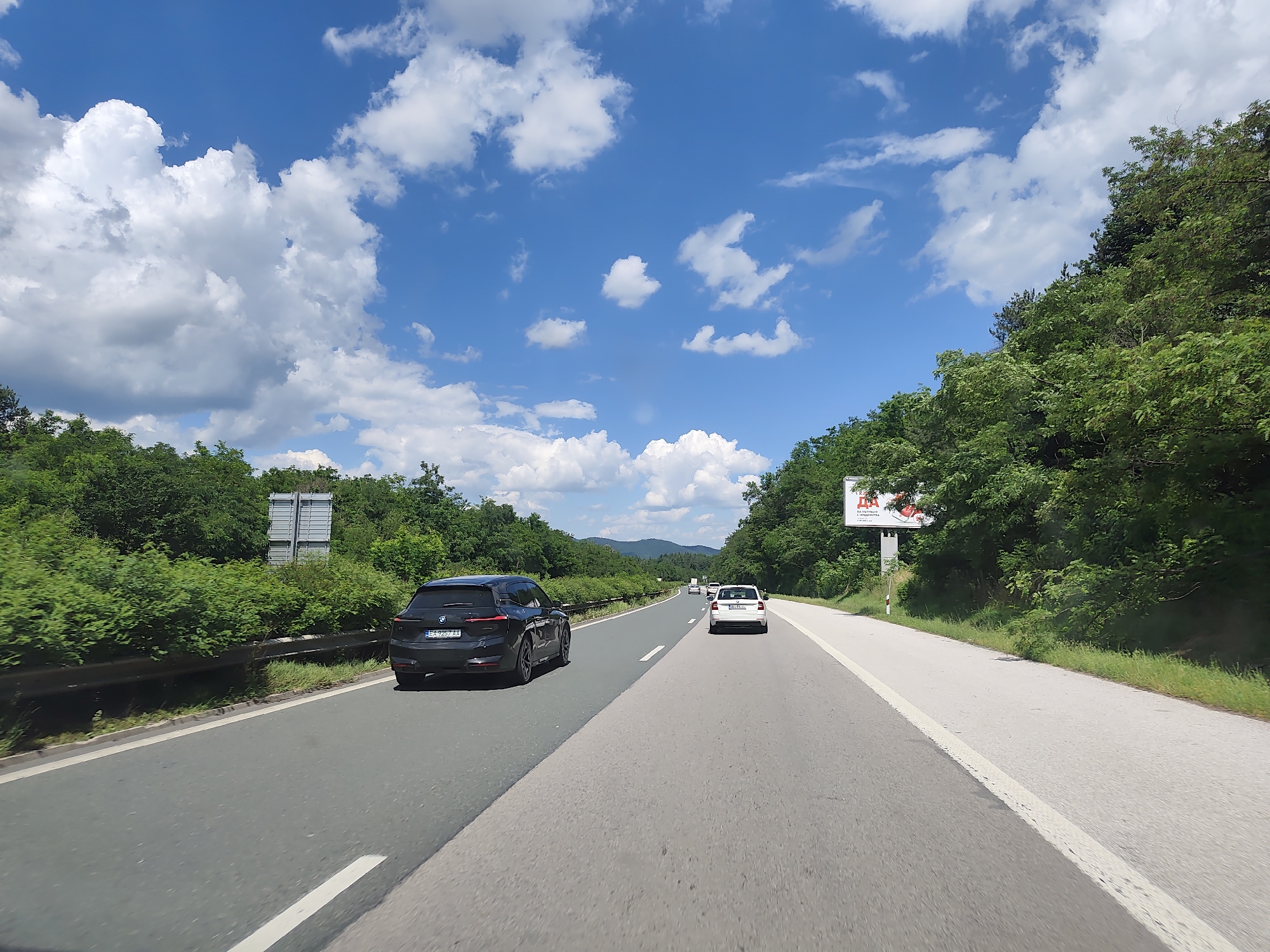 The image captures a view from a vehicle traveling on a highway, likely in or near Trudovets, Bulgaria, given the context of the prompt. The road is paved and appears to be a multi-lane highway with a clear division between lanes. In the foreground, the road surface is a mix of dark asphalt and lighter grey concrete. A white lane marker is visible on the right side of the road.
Several vehicles are present on the highway. To the left, a black SUV, possibly a BMW based on the visible license plate, is in the left lane. Further ahead, a white car is in the right lane. In the distance, other vehicles can be seen, suggesting moderate traffic.
The environment is characterized by lush green vegetation lining both sides of the highway. Dense trees and bushes create a natural barrier along the roadside. In the background, rolling hills covered in foliage are visible under a bright blue sky dotted with fluffy white clouds. A billboard is positioned on the right side of the highway amidst the trees, with some text and images on it. A metal guardrail runs along the left side of the road. The overall impression is a scenic highway drive on a clear, sunny day.