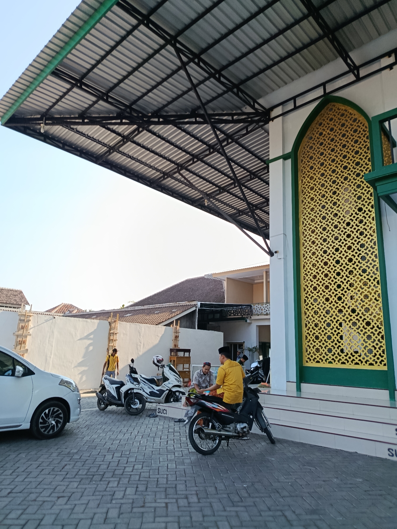 The image depicts the exterior of a mosque in Bondowoso, Indonesia. A large, corrugated metal roof with black metal framing extends from the building, providing shade over a paved courtyard. In the foreground, a white car is parked on the patterned brick pavement. Several motorcycles are also present, one of which is being sat on by a man wearing a yellow shirt and a black cap. Another man, wearing a checkered shirt and a white cap, is seated on a stool next to him, appearing to be in conversation. Behind them, a man in a yellow shirt and black cap stands near a construction area with wooden scaffolding against a white wall. The mosque itself features a large, arched window with intricate golden latticework and green trim. The sky is clear and bright.