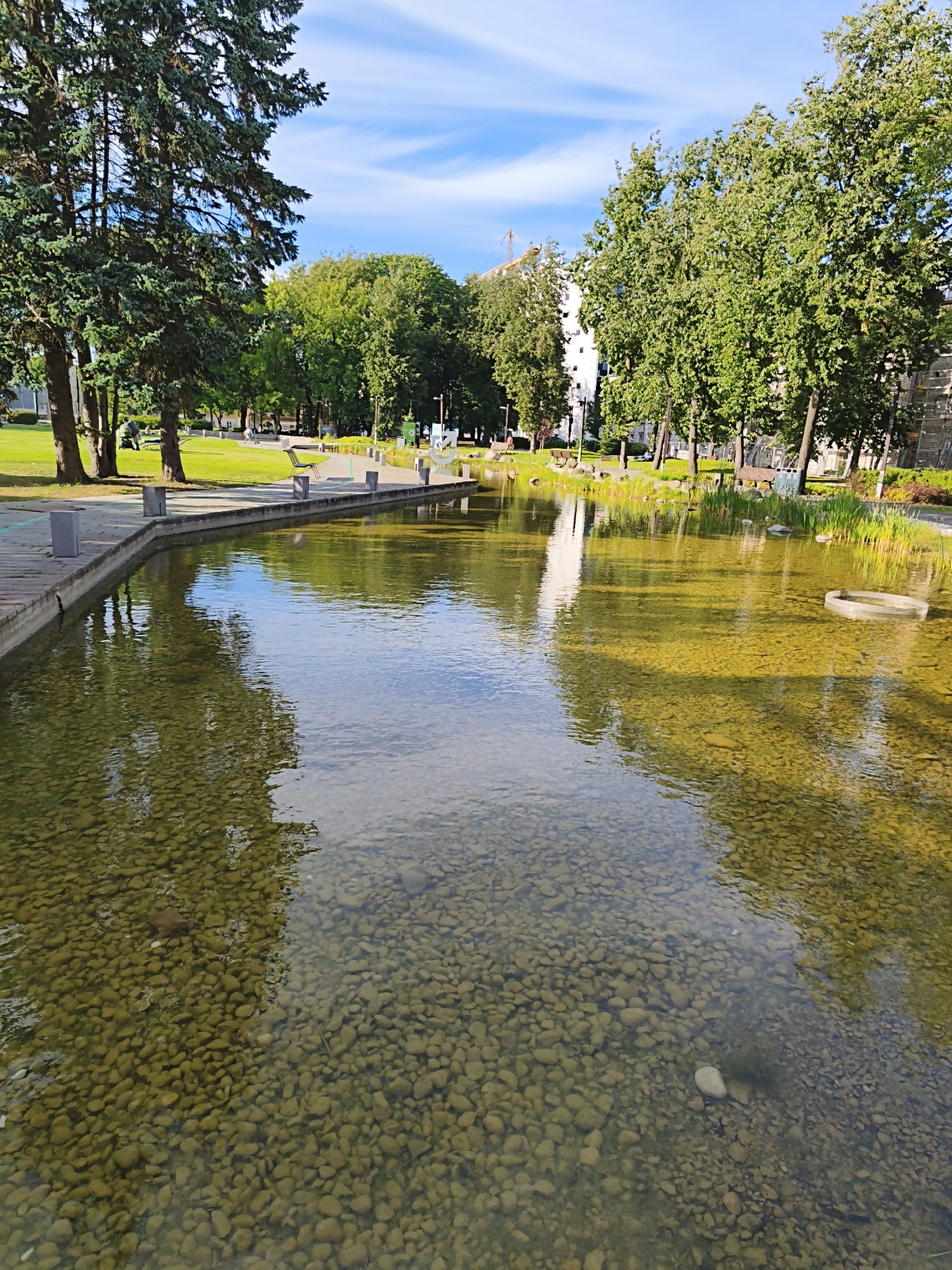 This is a low-angle outdoor photograph of a shallow, clear water feature in Tallinn, Estonia, likely in a park setting. The water is filled with small, smooth pebbles, visible through the transparent water. Sunlight filters through the trees, casting dappled reflections on the water's surface and illuminating the pebbles below with a golden hue.
To the left, tall evergreen trees with dense foliage frame the scene, their branches casting shadows. Beyond the water, a paved walkway runs parallel to its edge, bordered by square concrete posts. Further back, a grassy area stretches out, dotted with mature deciduous trees exhibiting lush green leaves. Several wooden benches are visible on the grass, with a few individuals scattered throughout the park, some appearing to be sitting or walking. In the background, a tall, modern white building stands behind the trees, with a construction crane visible above it. The sky is a clear blue, with wispy white clouds. On the right side of the water, reeds and aquatic plants grow along the edge. A circular, transparent object, possibly part of the water feature's design, is partially submerged in the water. The overall impression is one of a serene, well-maintained urban park on a sunny day.