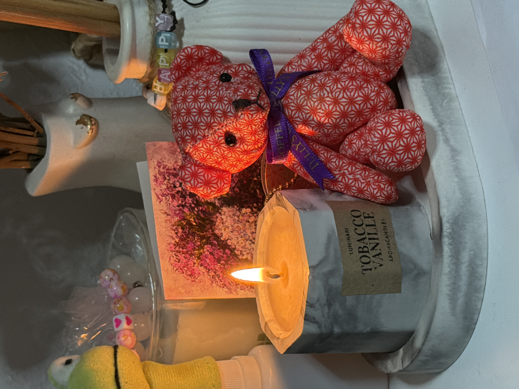 This is a close-up, eye-level, indoor photograph of a candle and a stuffed bear. The candle is in a hexagonal grey and white marbled concrete holder with a brown label that reads "LUMINARI TOBACCO VANILLE AROMACANDLE". The candle is lit, and the flame is a bright orange-yellow. To the right of the candle is a small red stuffed bear with a white geometric pattern. The bear is wearing a purple ribbon tied in a bow around its neck, which reads "TULLY'S COFFEE". Behind the bear and candle are various other objects, including a small pink floral greeting card, a white vase with dried reeds, and a smaller white vase with letters spelling "HAPPY". A bright green stuffed frog toy is visible on the far left of the image. The lighting suggests it is nighttime or the room is dimly lit. There are no people present in the image, and no elements suggest a specific location in Moscow, Russia.