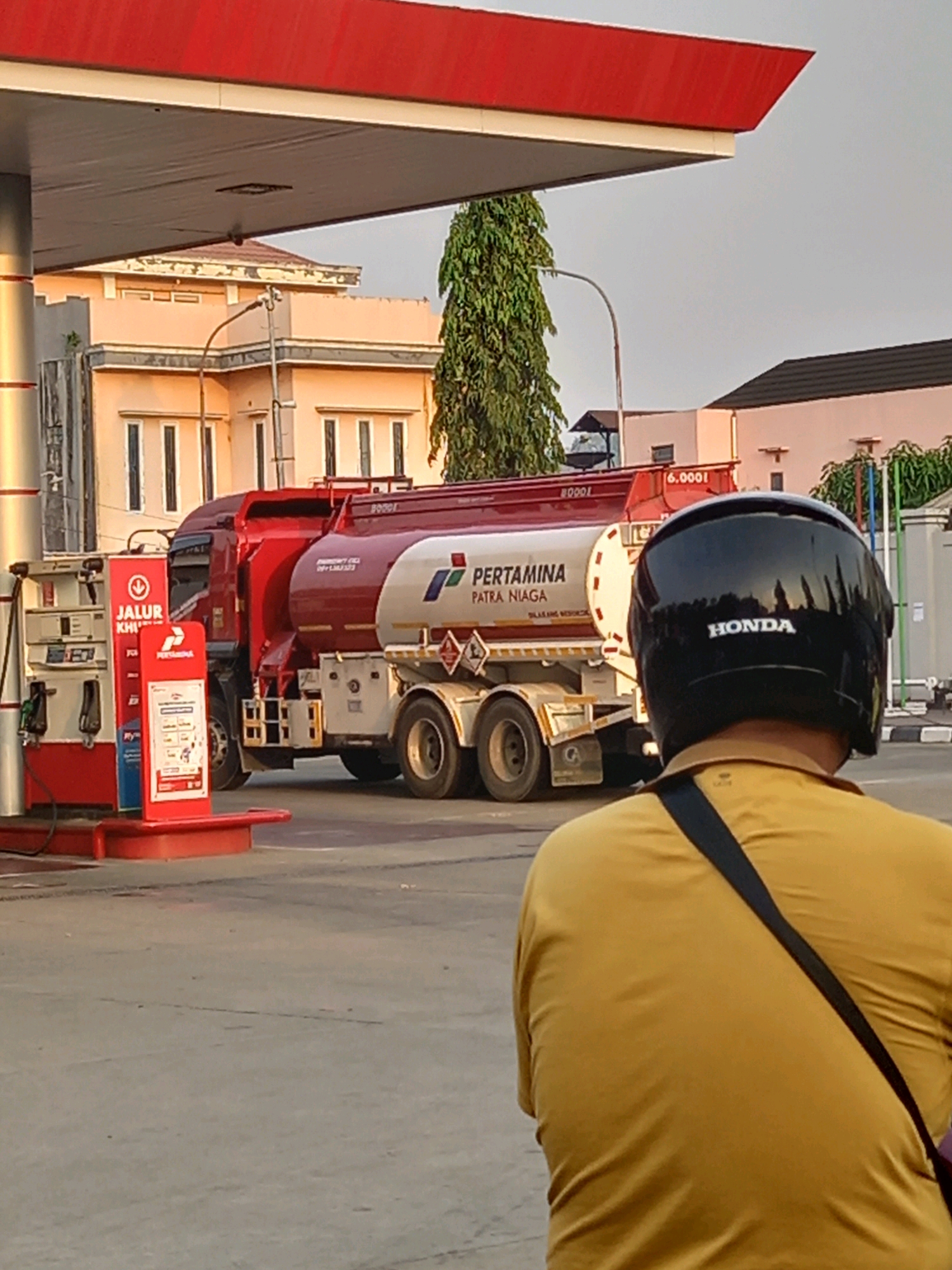 The image depicts a Pertamina gas station in Samarinda, Indonesia, with a tanker truck delivering fuel. The perspective is from behind a person wearing a yellow shirt and a black helmet with "HONDA" written on the back, seated on a vehicle, observing the scene. The gas station canopy, painted red and white, extends over the pumps. A red and white tanker truck, labeled "PERTAMINA PATRA NIAGA," is parked at the station, its large cylindrical tanks visible. Various gas pumps, with digital displays and hoses, are to the left of the truck. In the background, there are buildings with light-colored walls and dark roofs, and a tall, green tree. The overall lighting suggests it is either early morning or late afternoon.
