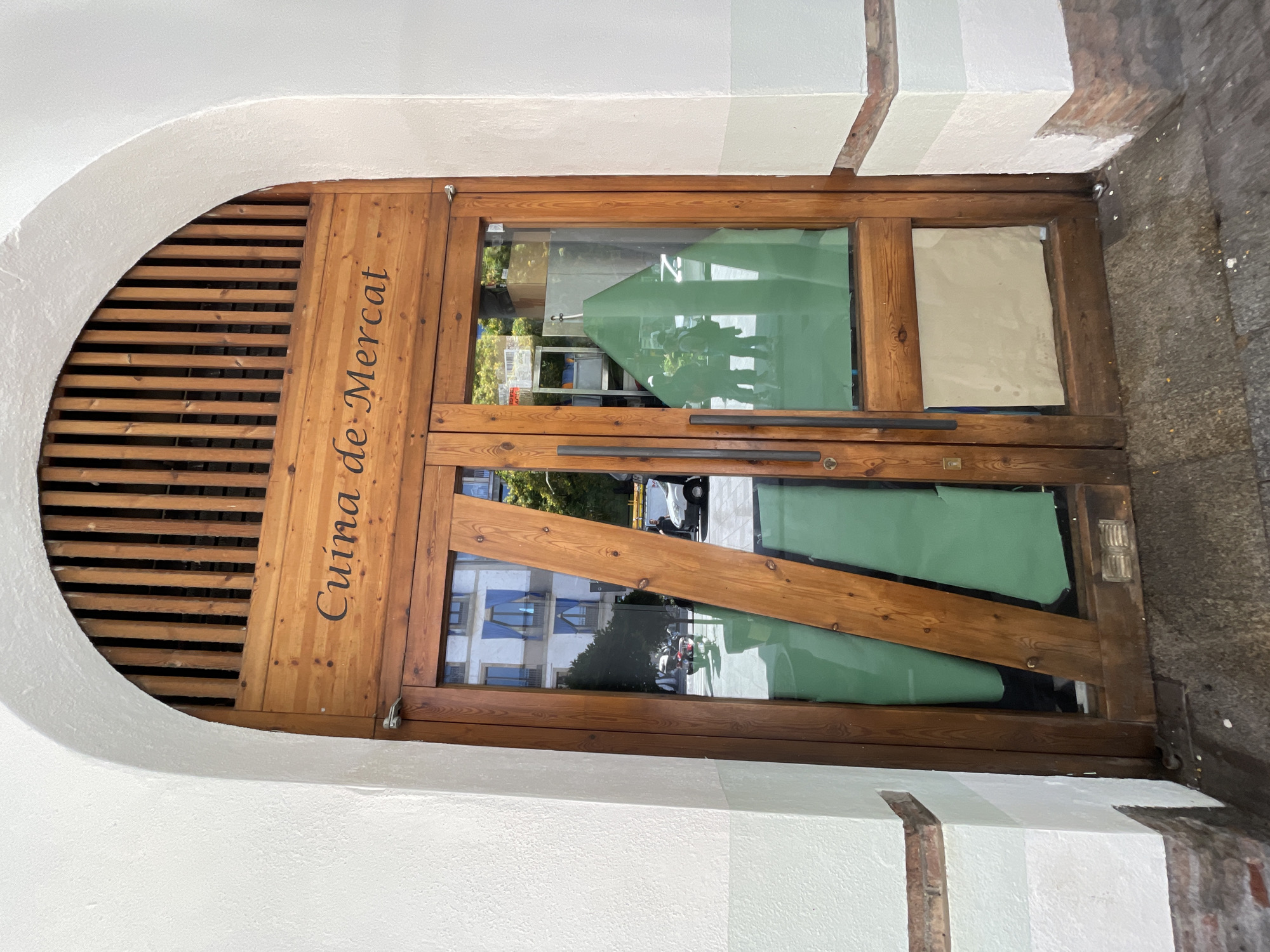 An arched wooden double door, with a slatted wooden transom above, serves as the entrance to a establishment in the city of Barcelona, Spain. The door is set within a white textured stucco archway, which transitions to a light green/grey painted band and exposed brickwork at the base. The door features the phrase "Cuina de Mercat" in black script on its upper wooden panel. Both door leaves contain large glass panels. The left door's glass is partially obscured from the interior by two diagonal wooden cross-pieces and green material, forming triangular shapes. The right door's glass is also partially covered with green material, and the lower portion of its glass panel is covered with a beige sheet from the interior. Dark, vertical bar handles are present on the right door. The glass reflects an urban street scene, including a white building with blue window coverings, a white van, a motorcycle, several pedestrians, and green trees under a bright sky. The ground directly in front of the door is dark grey paving with scattered white debris.