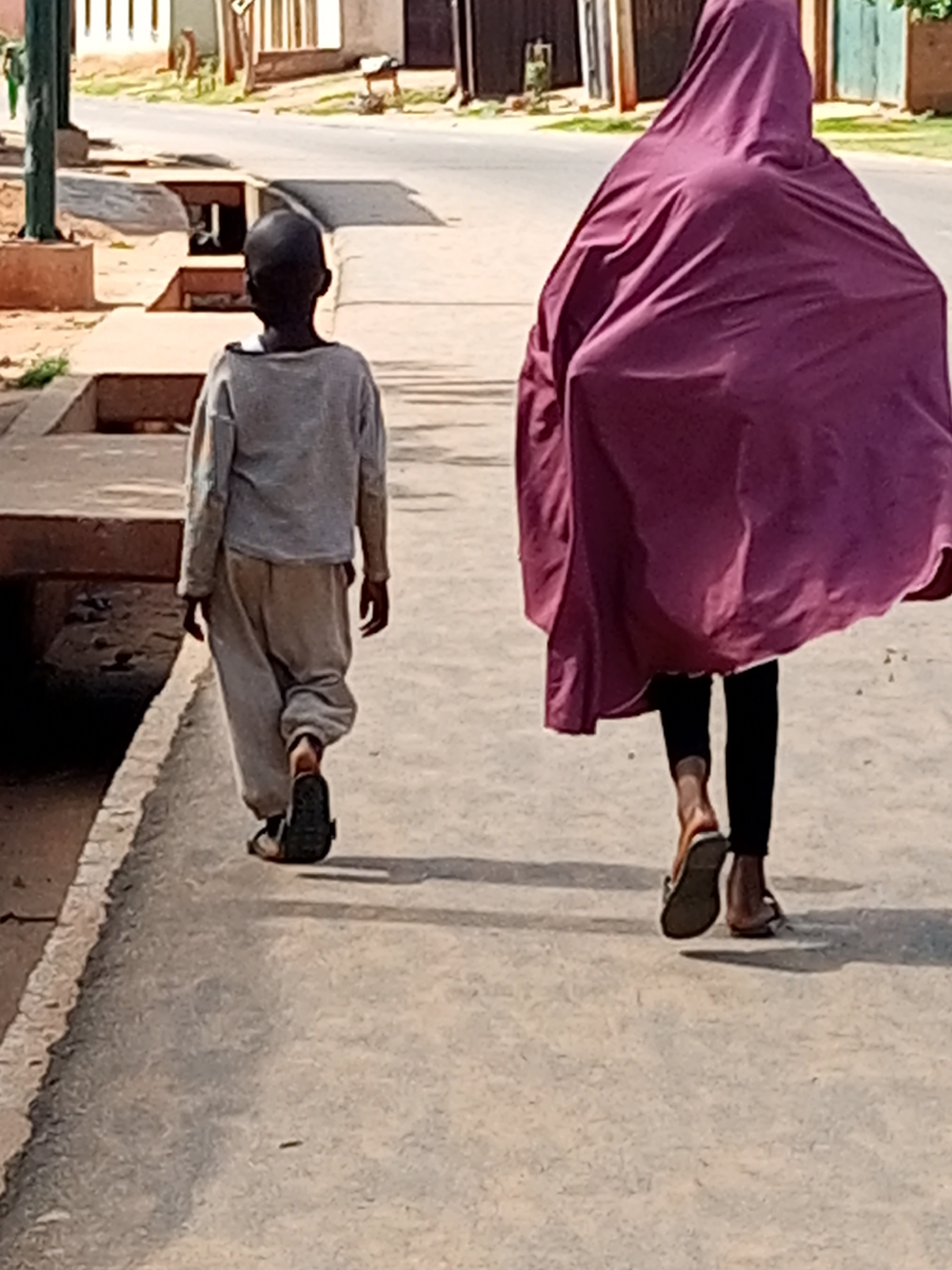 In Gombe, Nigeria, two individuals walk on a paved sidewalk adjacent to a street. On the left, a young person, appearing to be a child, walks away from the camera. They are wearing a light grey long-sleeved shirt and loose-fitting grey pants, with one foot shod in a dark sandal. To the right, another individual, whose gender is obscured by their attire, walks away from the camera, facing the same direction as the child. This person is covered in a flowing, deep pink or magenta fabric, which drapes over their body and extends downwards. They are wearing black leggings and open-toed sandals. The sidewalk is bordered by a shallow concrete ditch or drainage channel on the left. The background shows a street with vehicles and buildings lining it, typical of an urban environment in Gombe. The lighting suggests a sunny day, casting shadows on the ground.