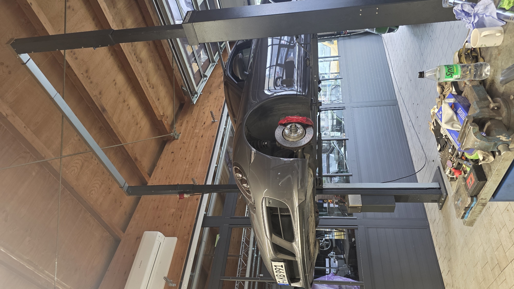 A gray Porsche is elevated on a hydraulic car lift in an indoor garage. The car's front left wheel is exposed, revealing a red brake caliper and disc. The license plate reads "F KB 991". Above the car, a wooden ceiling is visible with exposed beams and electrical conduits. To the left, a white air conditioning unit is mounted on the ceiling. In the background, a row of gray garage doors are closed. In the foreground, a workbench is covered with various tools, including a vise, gloves, and a bottle of Sprite. The floor is tiled with light gray and beige squares. The scene appears to be a service bay in an automotive repair shop. There are no people visible in the image.
